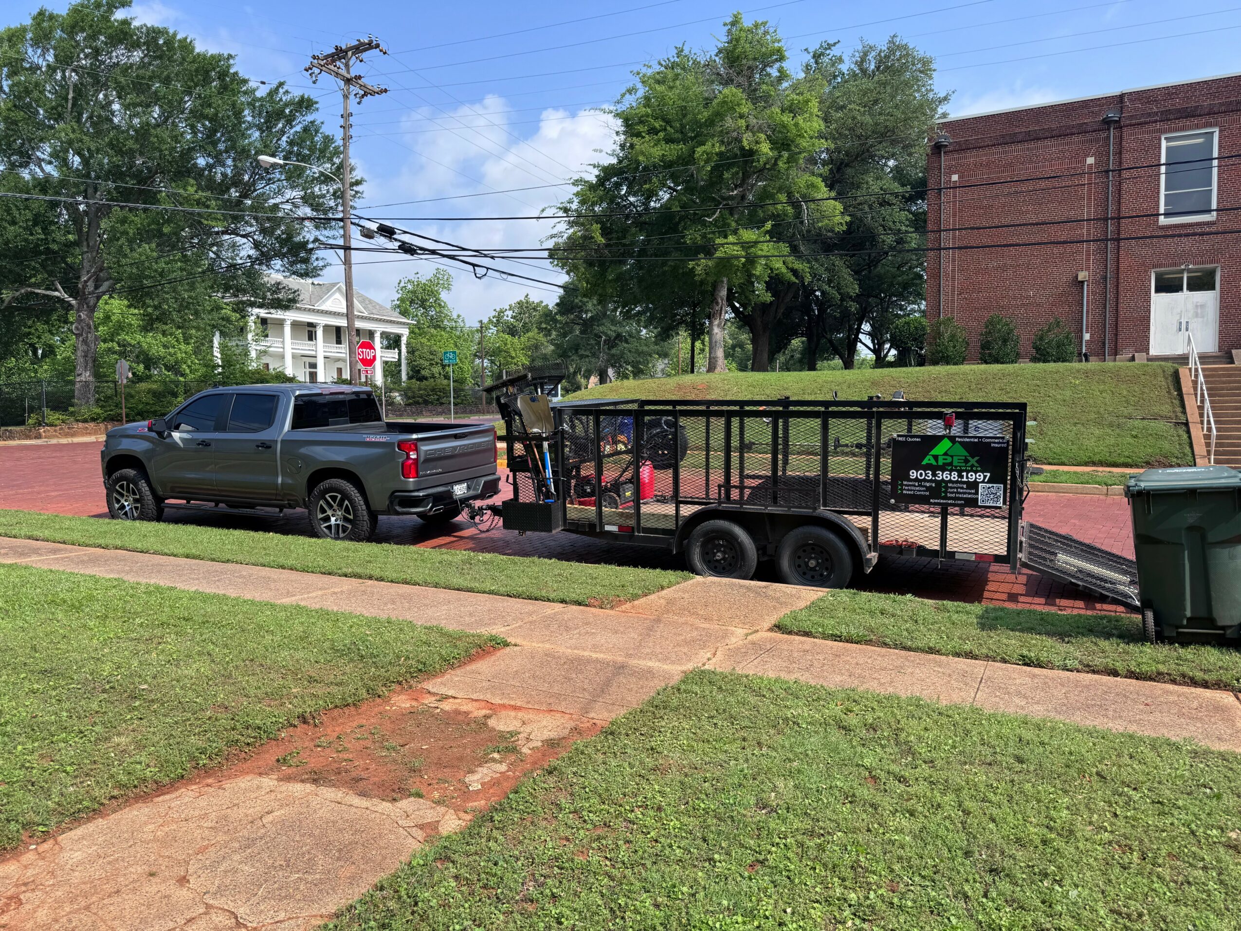 Black pickup truck with a trailer attached parked on a brick driveway, with trees and a brick building in the background.