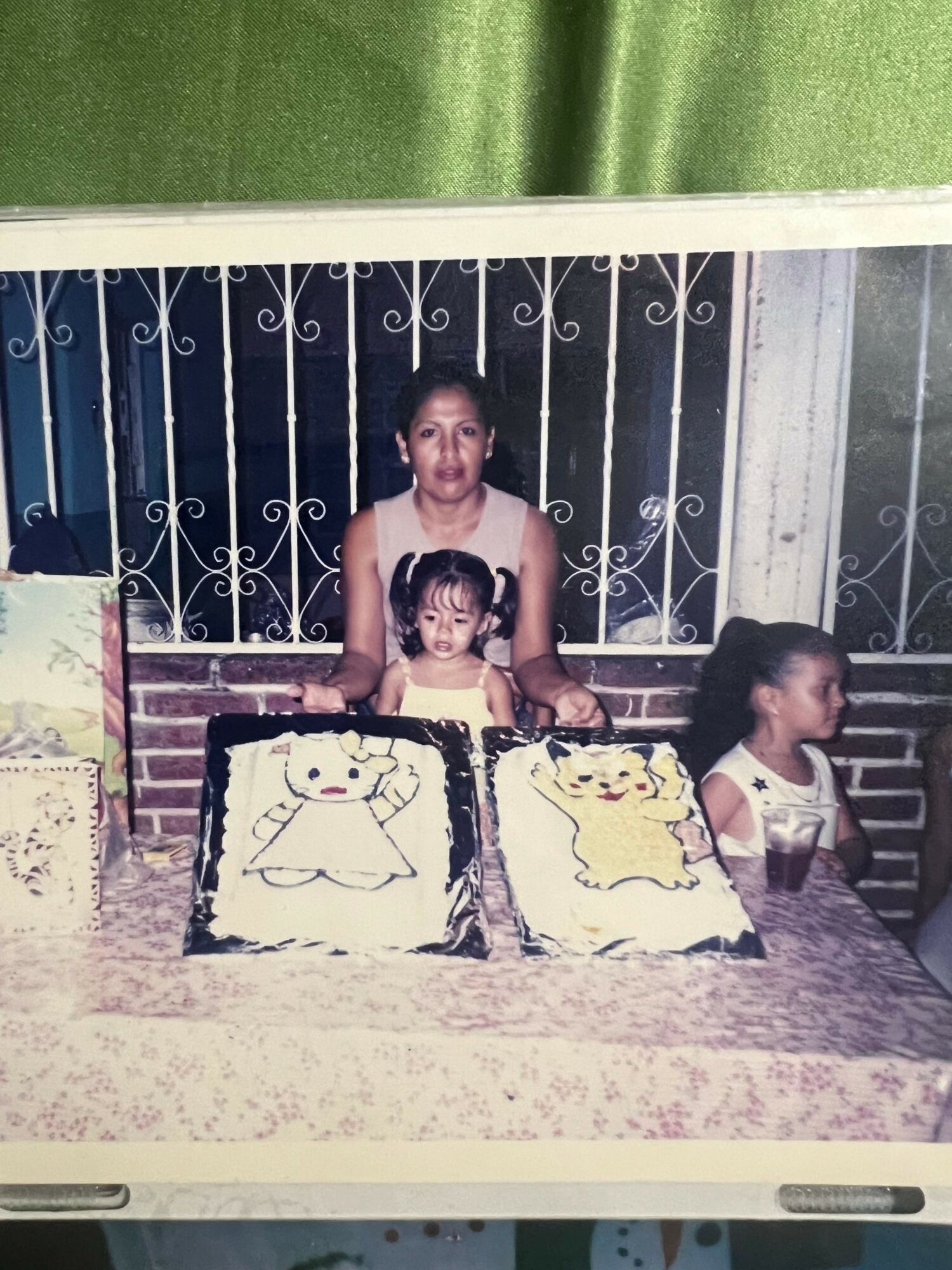 Three children around a table with a cake, one girl is blowing out candles, two others watch, background shows a gate.