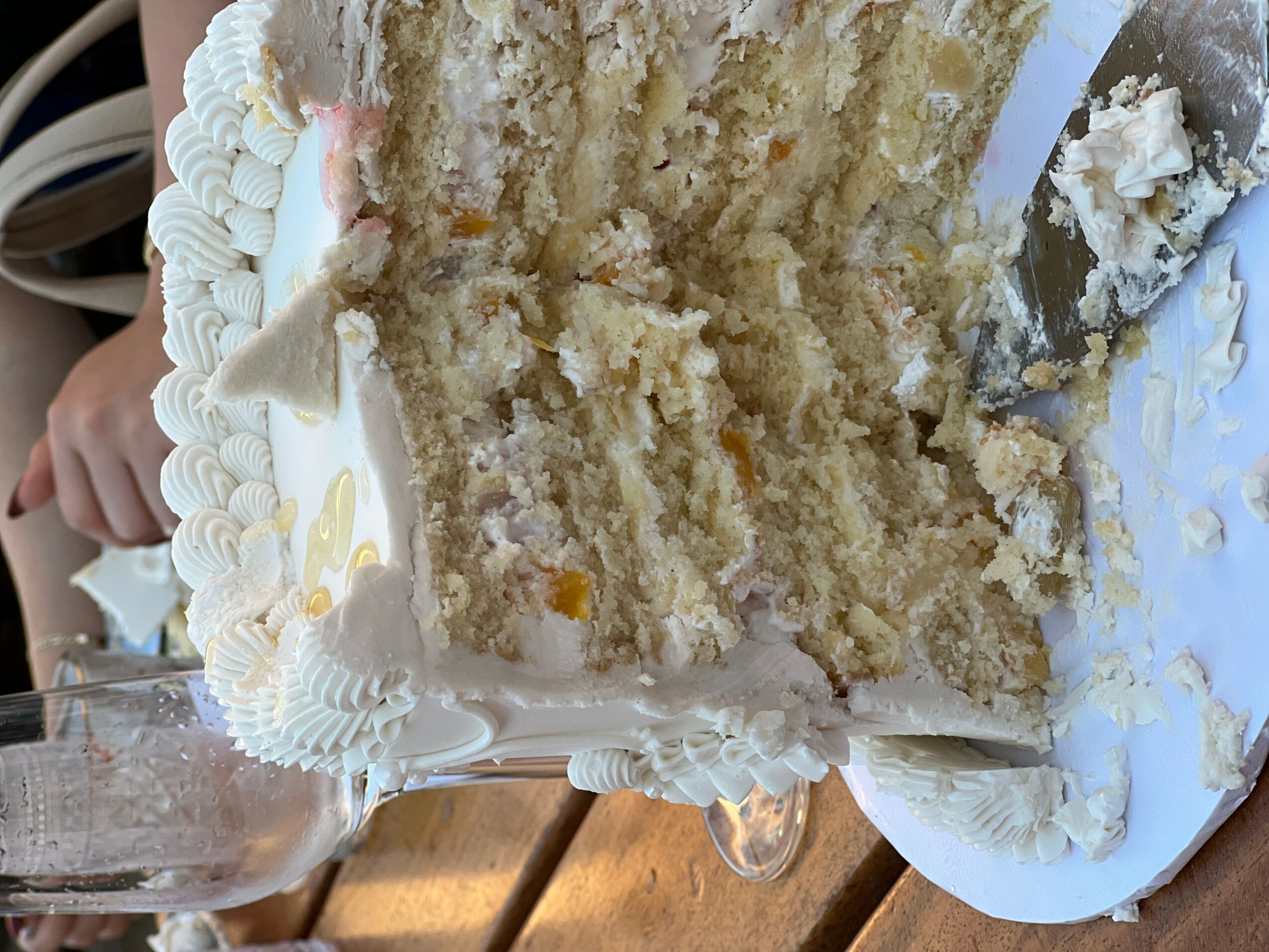 A partially eaten layered cake with white frosting and decorative piping on a white plate, with a person holding a glass nearby.