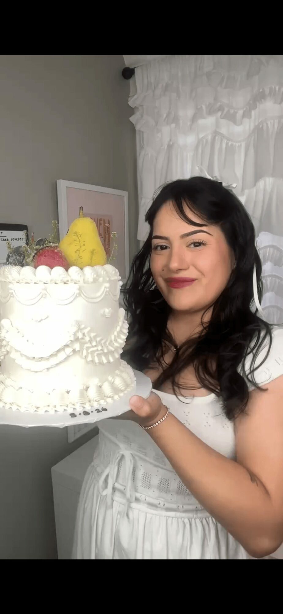 Woman with dark hair and red lipstick holding a decorated cake with yellow and pink decorations, smiling.
