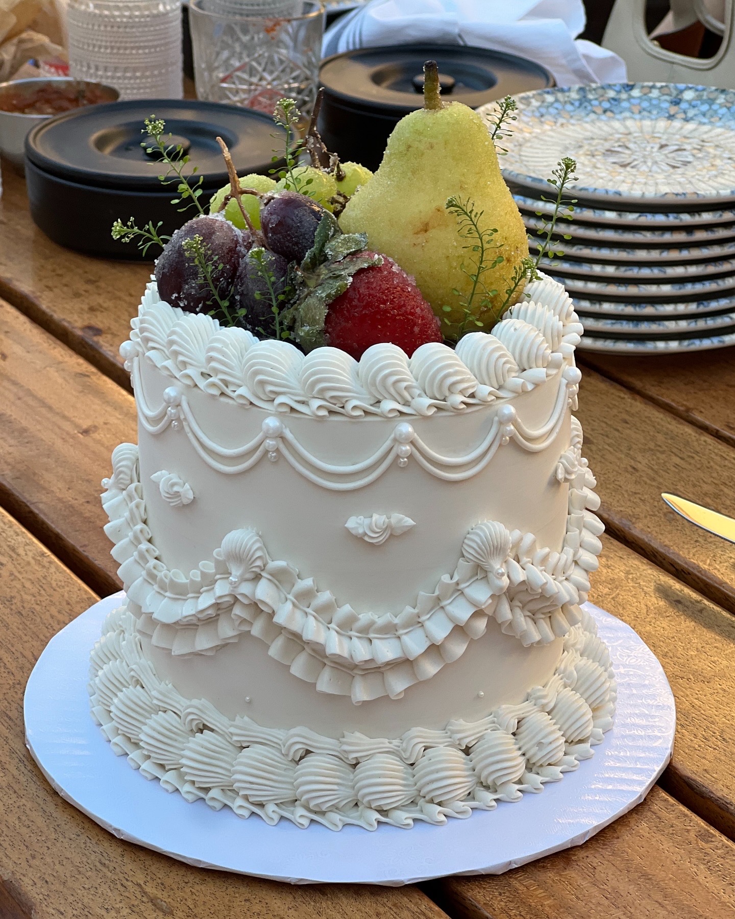 White decorated cake topped with a pear, grapes, and a strawberry, on a wooden table with plates and glasses in background.
