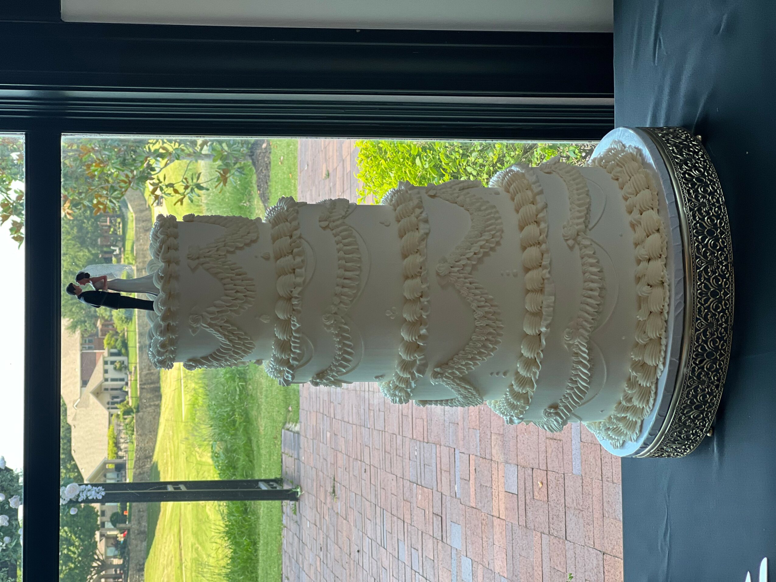 Tall, multi-tiered white wedding cake with decorative icing, on a round base, near a window with outdoor view.