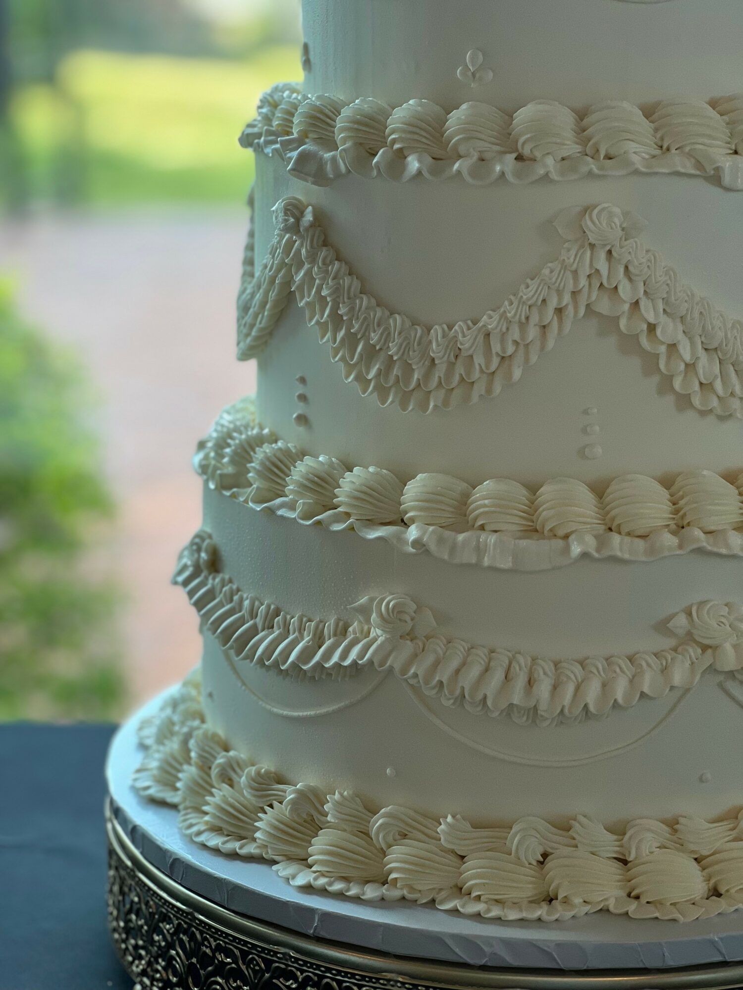 Close-up of a multi-tiered white cake with decorative icing and piping, outdoors with blurred greenery background.