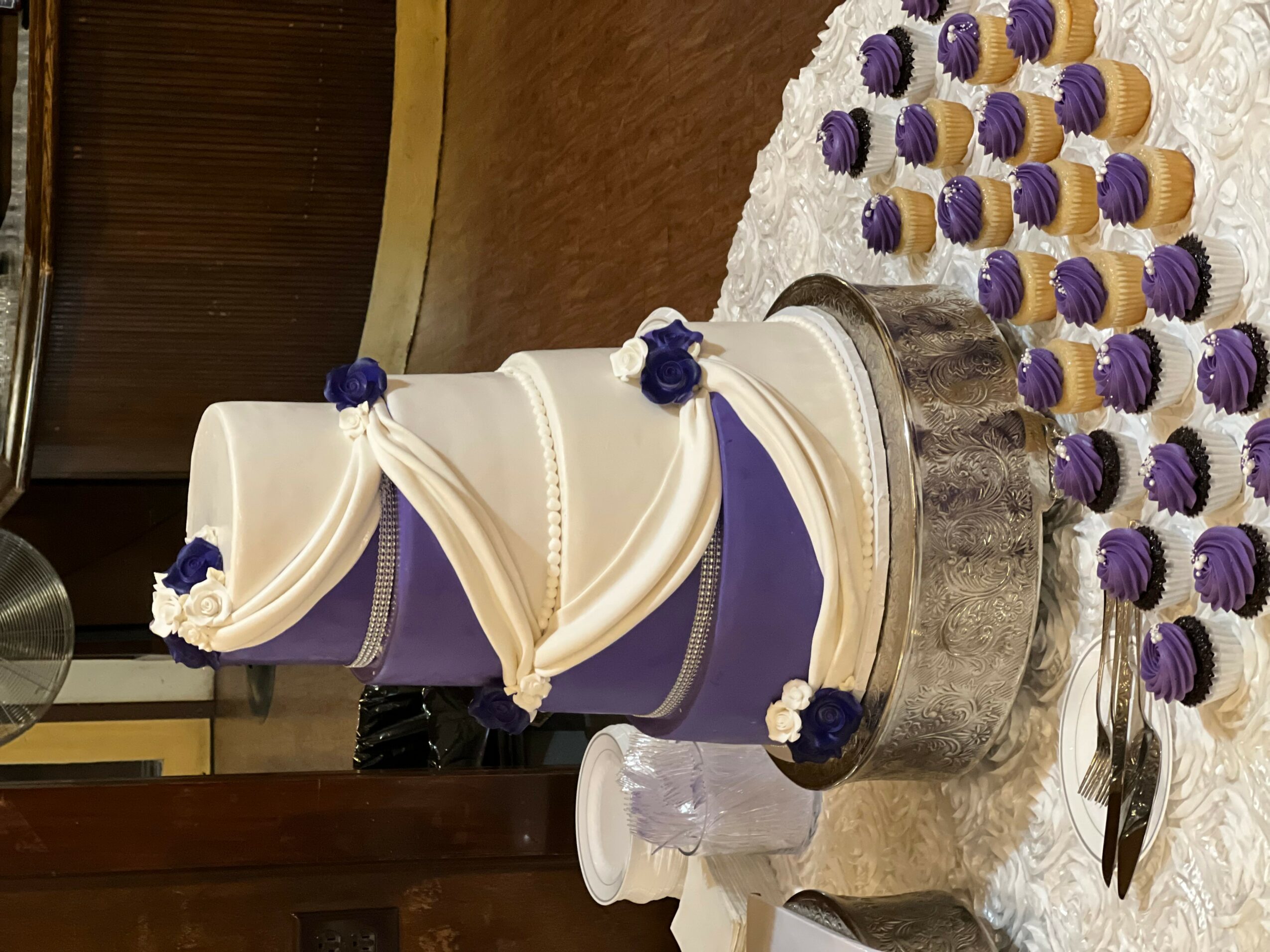 Tall, multi-tiered wedding cake with white and purple decorations, surrounded by purple and white cupcakes, on a table.