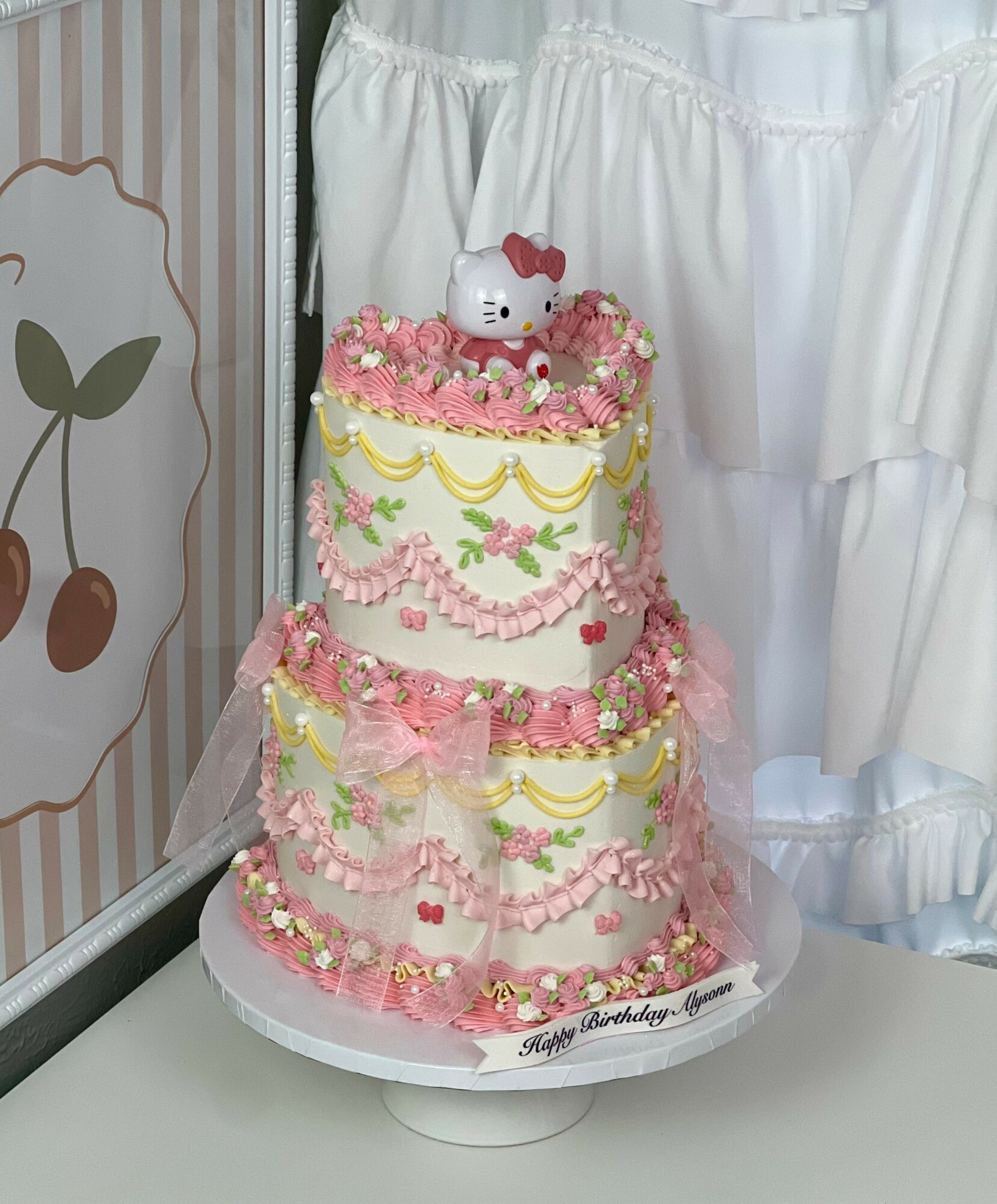 Two-tiered pink and white birthday cake with Hello Kitty topper, decorated with pink, yellow, and green icing, and small flowers.