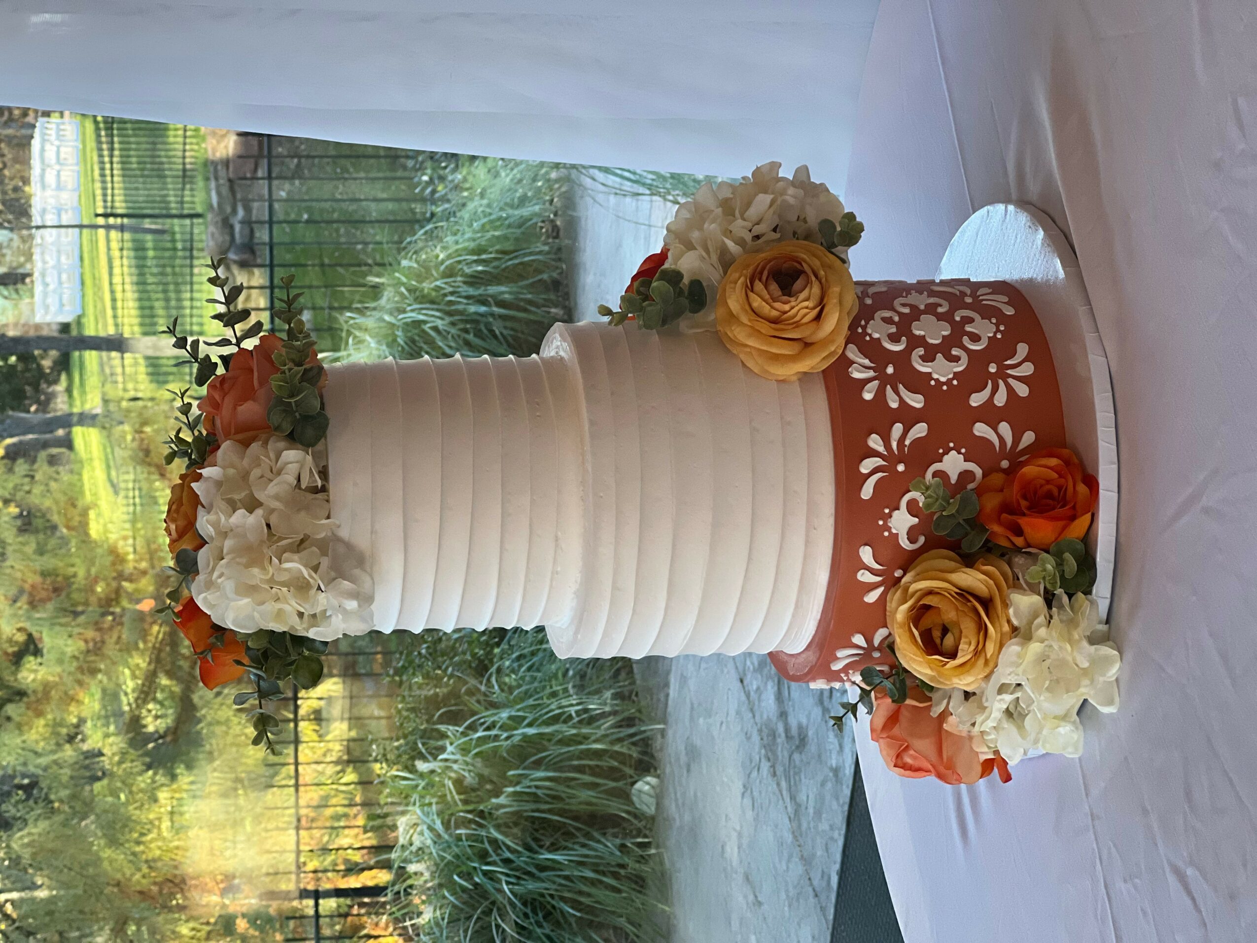 A tall white tiered cake with floral decorations, placed on a table near a window with greenery outside.