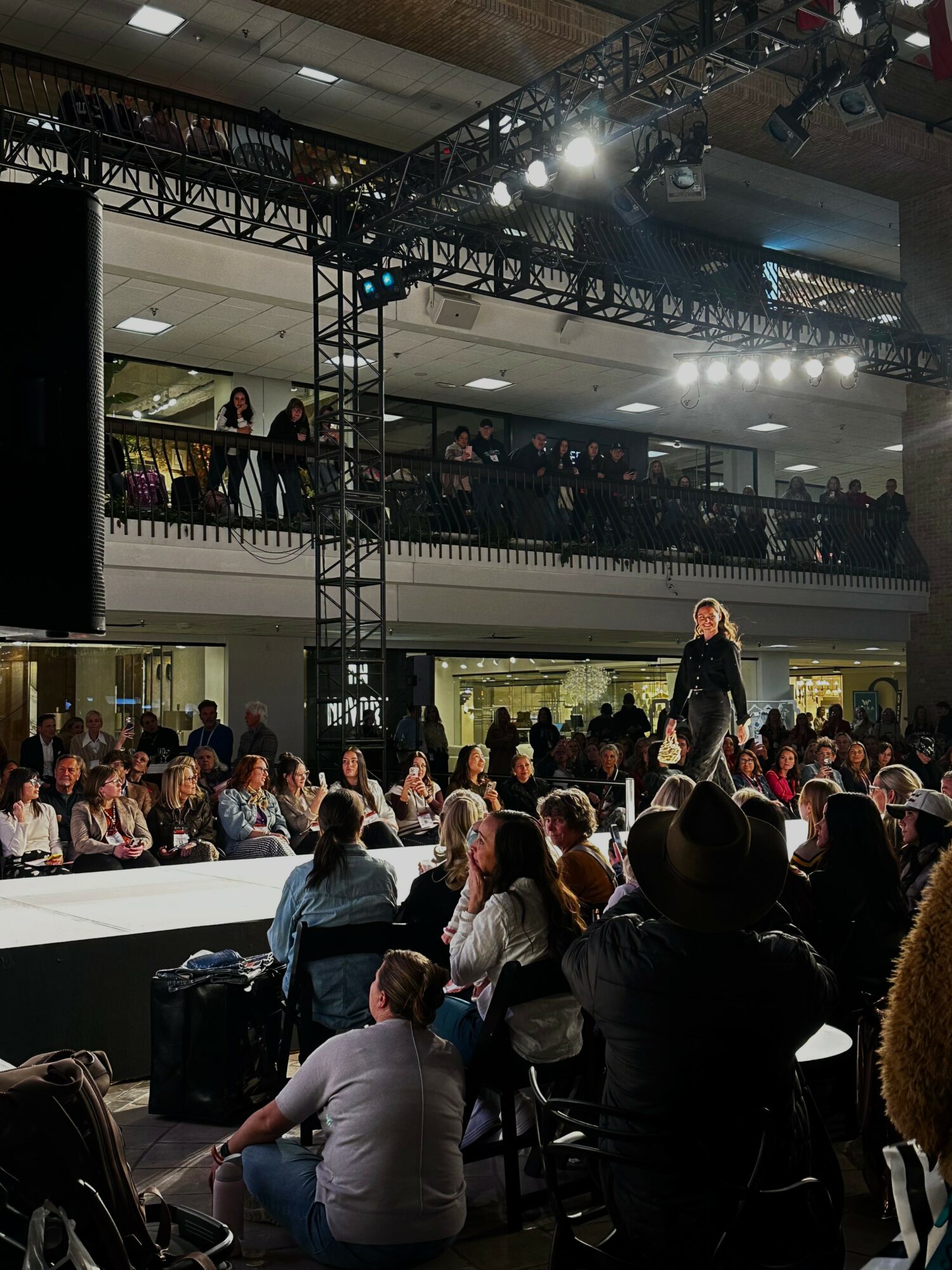 Fashion show runway with a model walking, audience seated and standing, two levels of spectators, bright lighting, indoor setting.