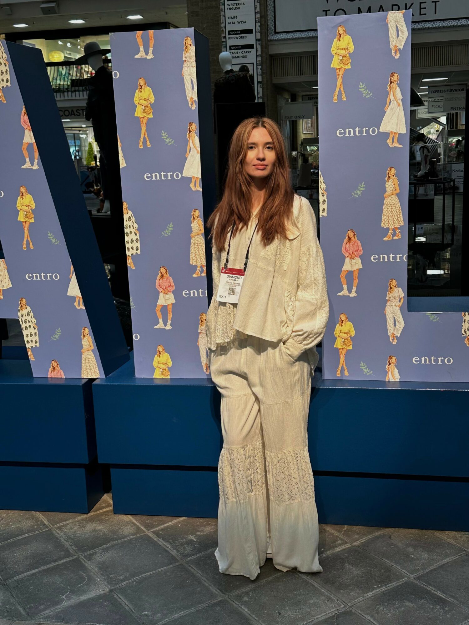 Woman with long hair in cream-colored outfit standing in front of a backdrop with fashion illustrations, indoors.