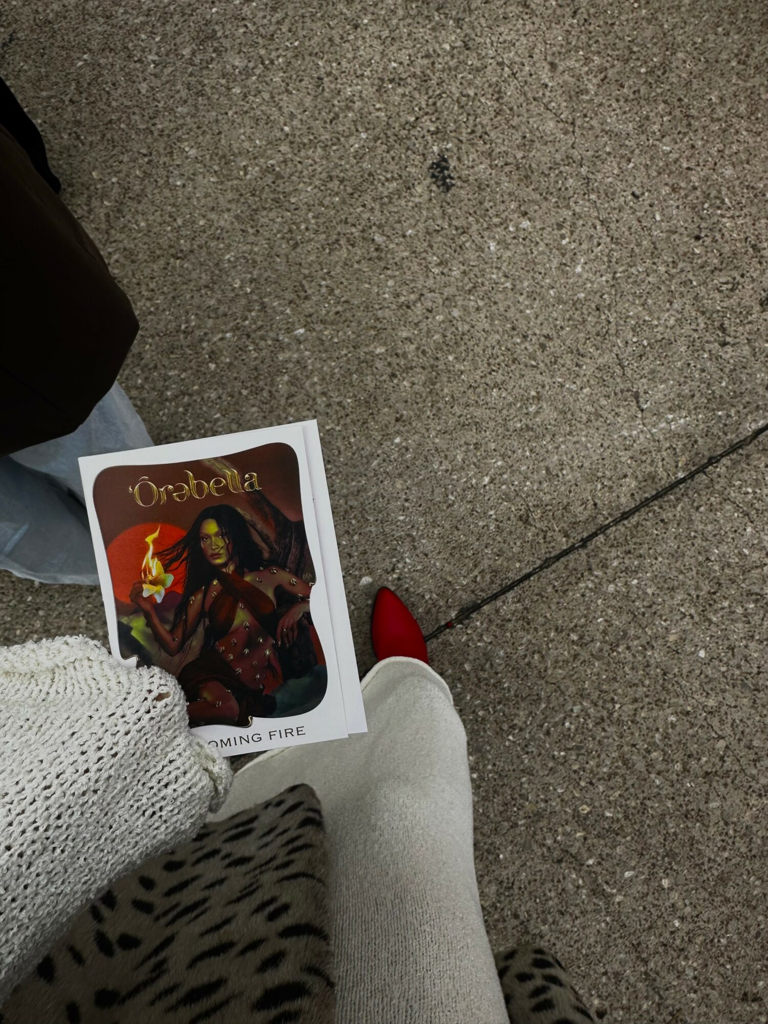 Person holding a card with a fiery character illustration, standing on a concrete sidewalk, wearing white pants and red shoes.