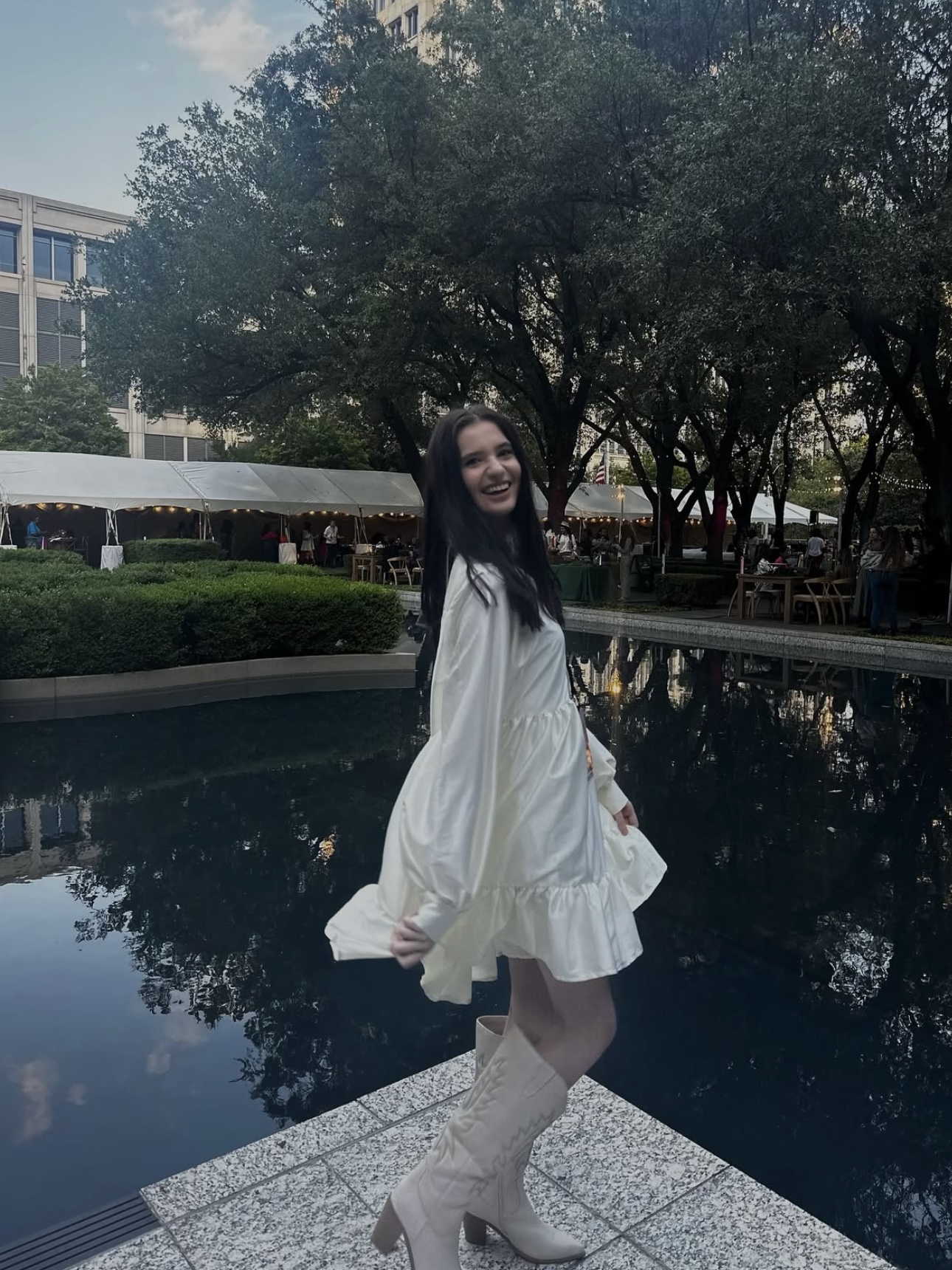 Young woman in white dress and knee-high boots standing by a pool with trees and buildings in background.