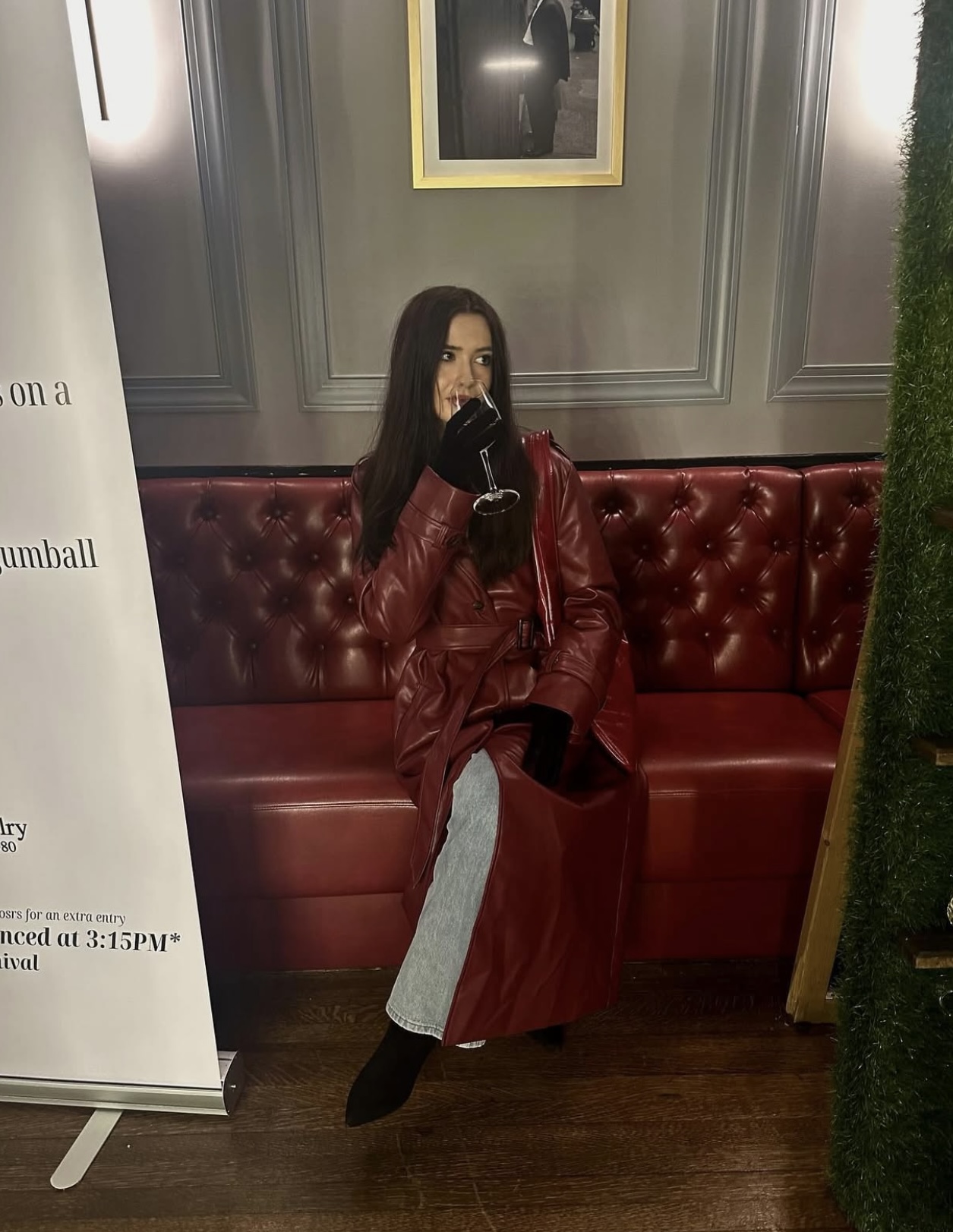 Woman sitting on a red leather bench, wearing a red jacket and gray pants, covering her mouth with her hand, in a decorated room.