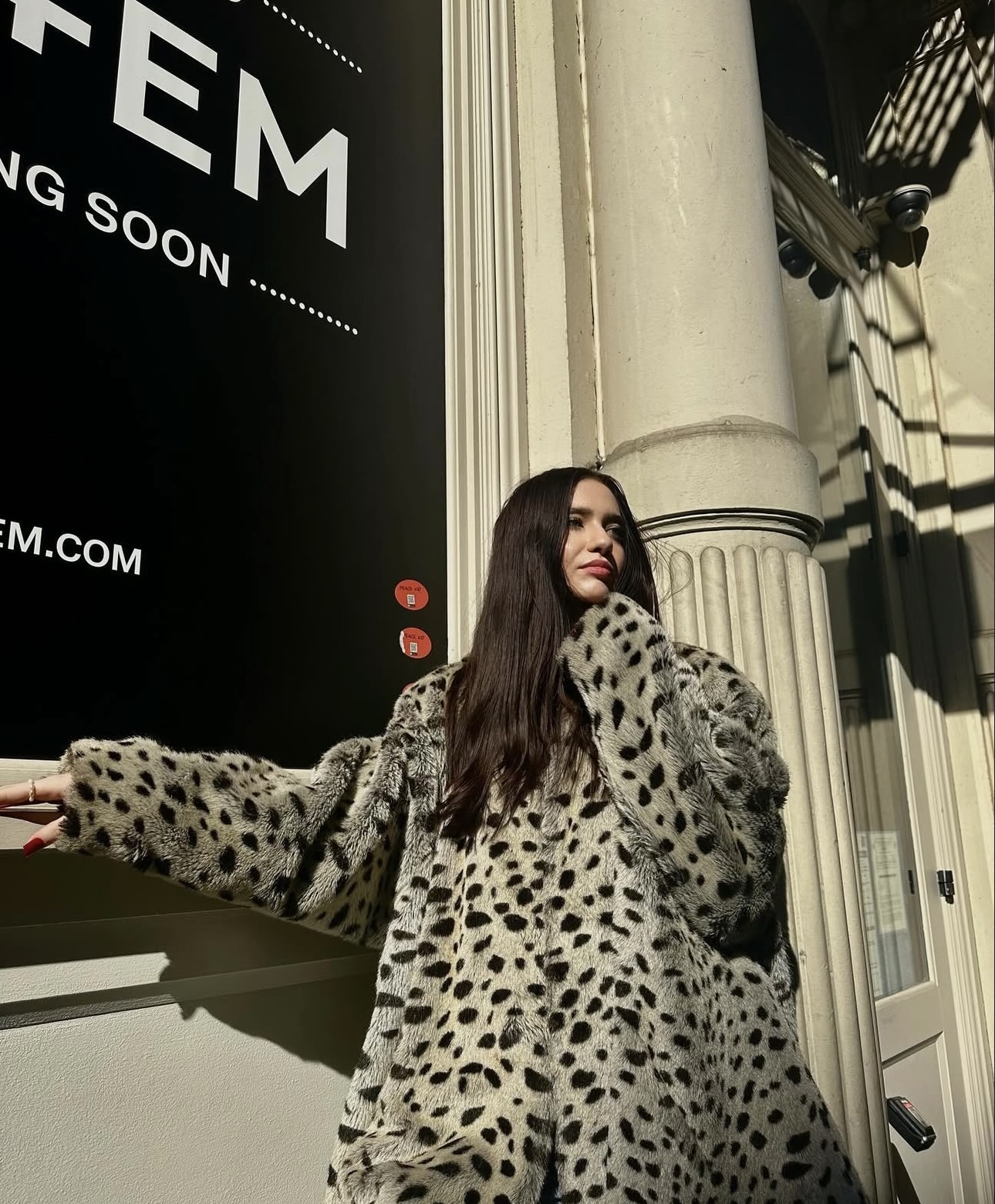Young woman with long dark hair wearing a spotted coat, standing next to a column outside a building, sunlight casting shadows.