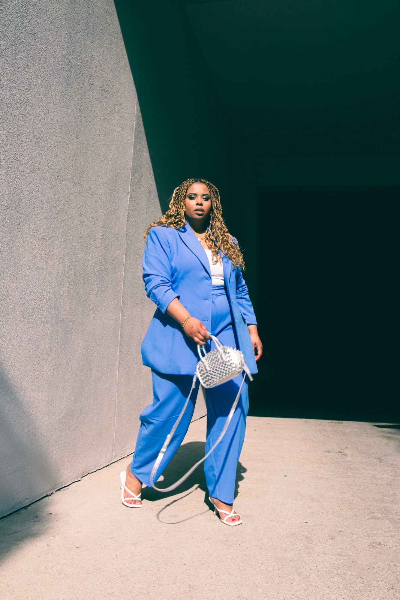 Woman in blue suit holding a white handbag, standing against a wall with a dark background.