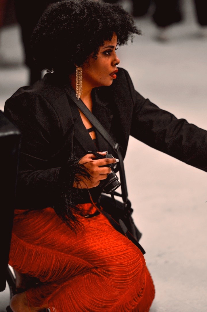 Woman with curly hair wearing a black jacket and red skirt, sitting outdoors, holding a camera, with a blurred background.