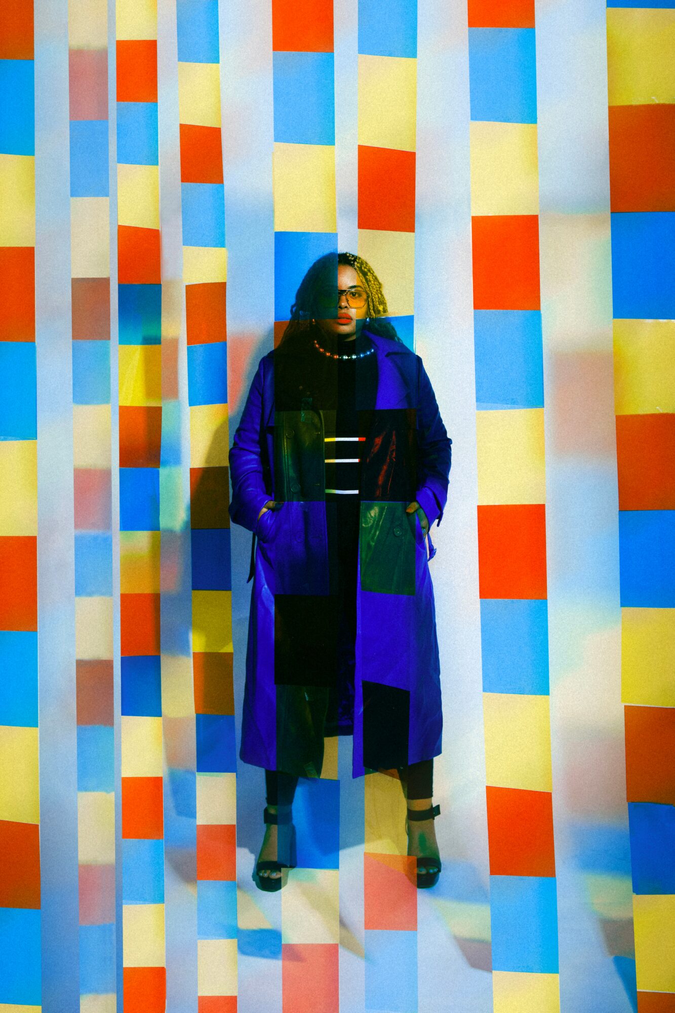 Person standing in front of a colorful, geometric patterned background with vertical stripes of squares in red, yellow, blue, and white.