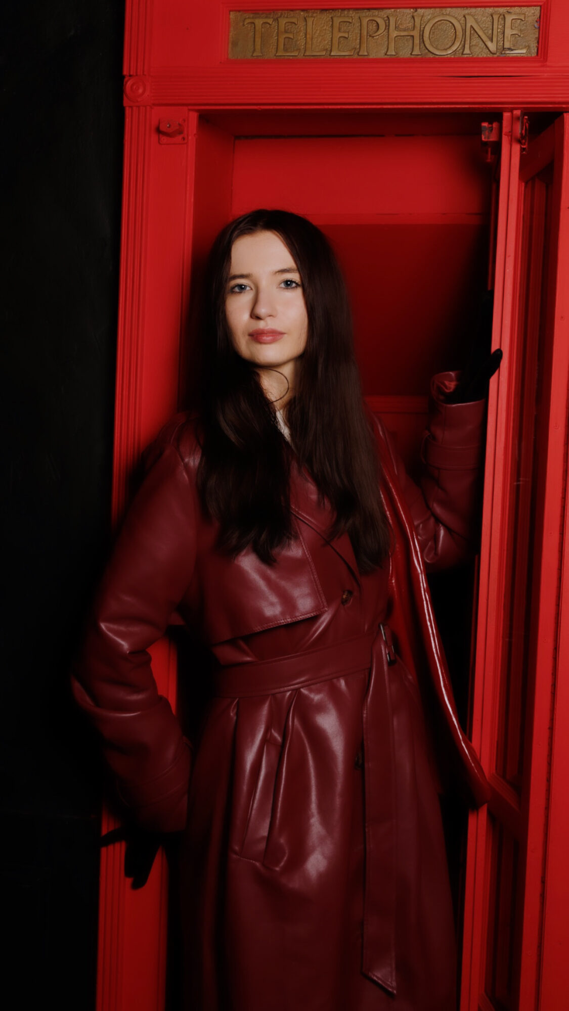 Young woman with long dark hair standing inside a red phone booth, wearing a maroon leather coat, holding the door handle.