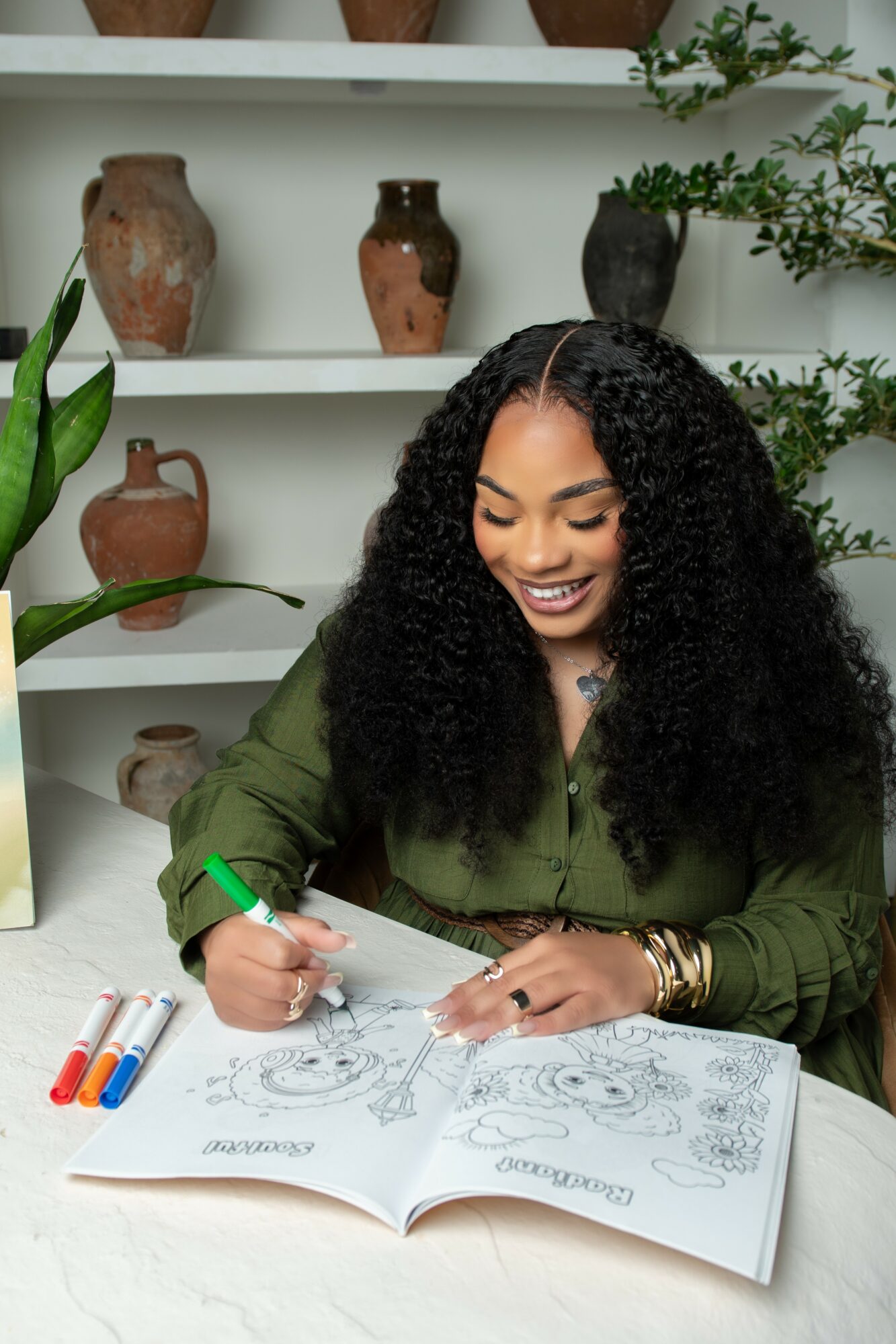 Woman with curly hair drawing in a sketchbook, smiling, surrounded by pottery and plants, wearing a green shirt.