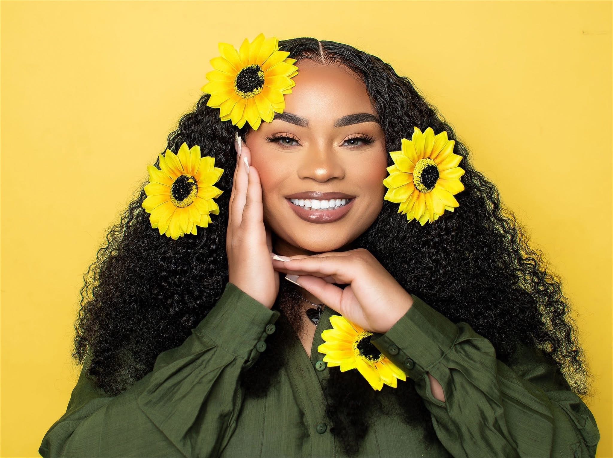 Smiling woman with curly hair and yellow flowers in her hair, wearing a green shirt, against a yellow background.