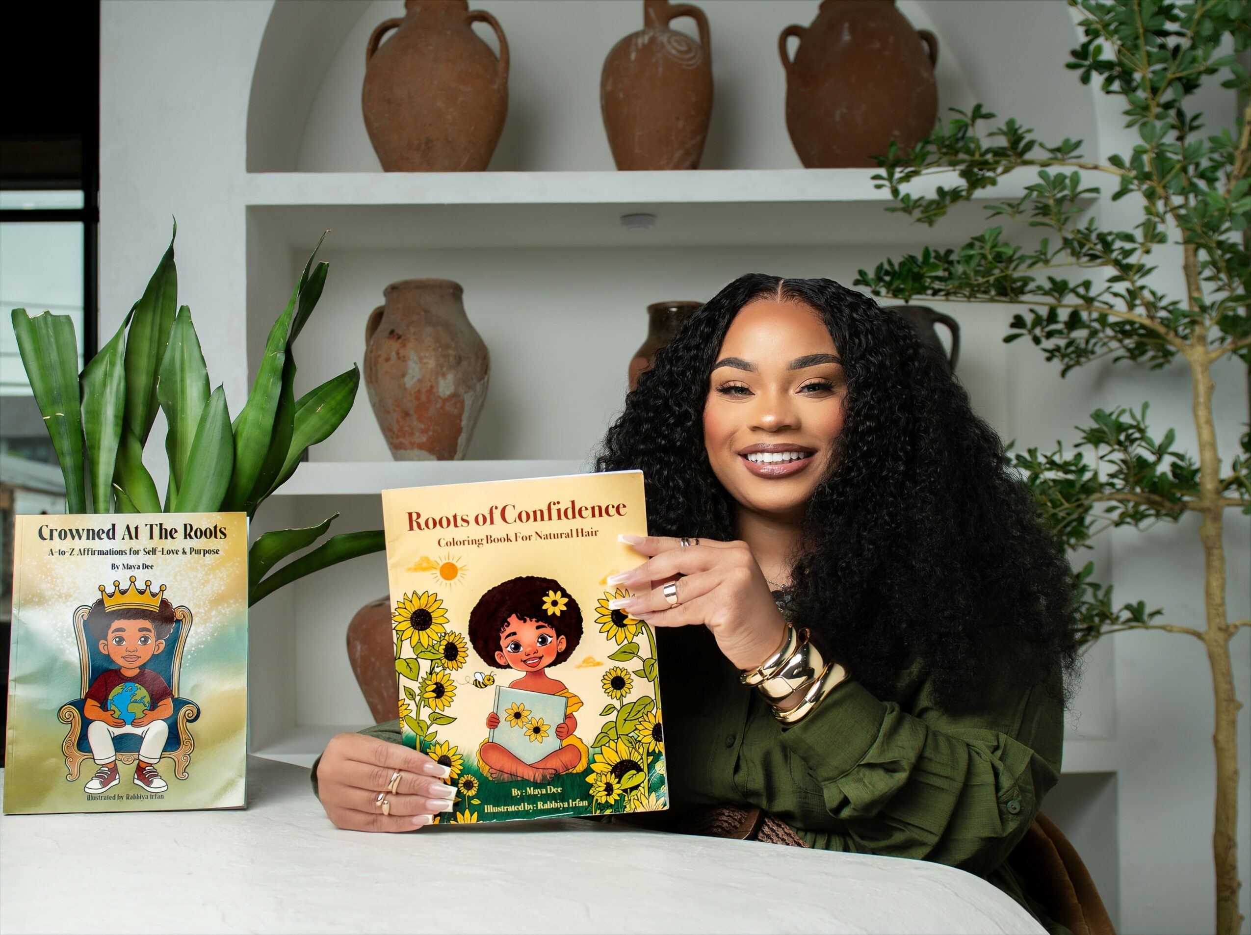 Smiling woman with curly hair holding a children's book, sitting at a table with plants and decorative vases behind her.