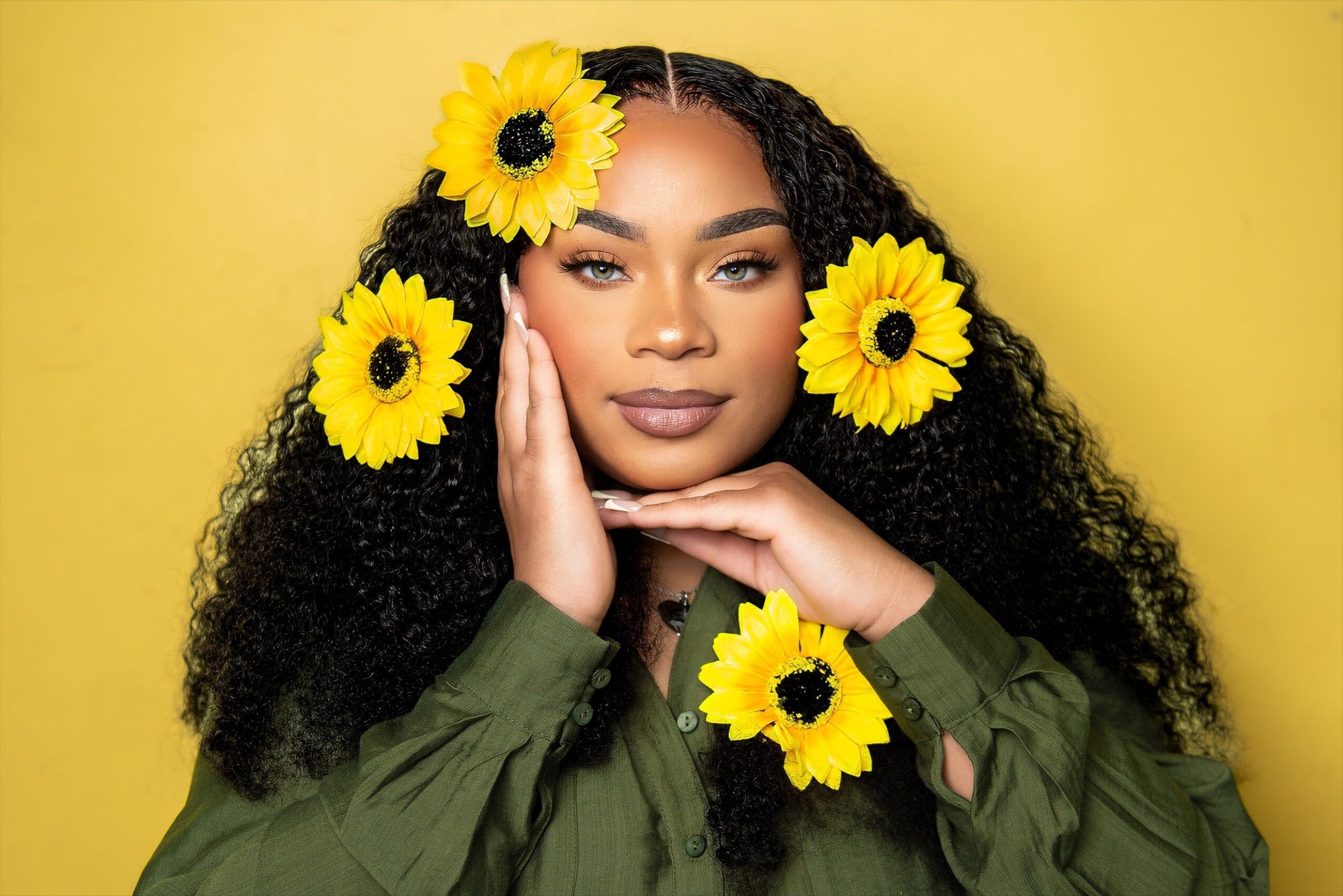 Woman with long curly hair and yellow flowers in her hair, wearing a green top, against a yellow background.