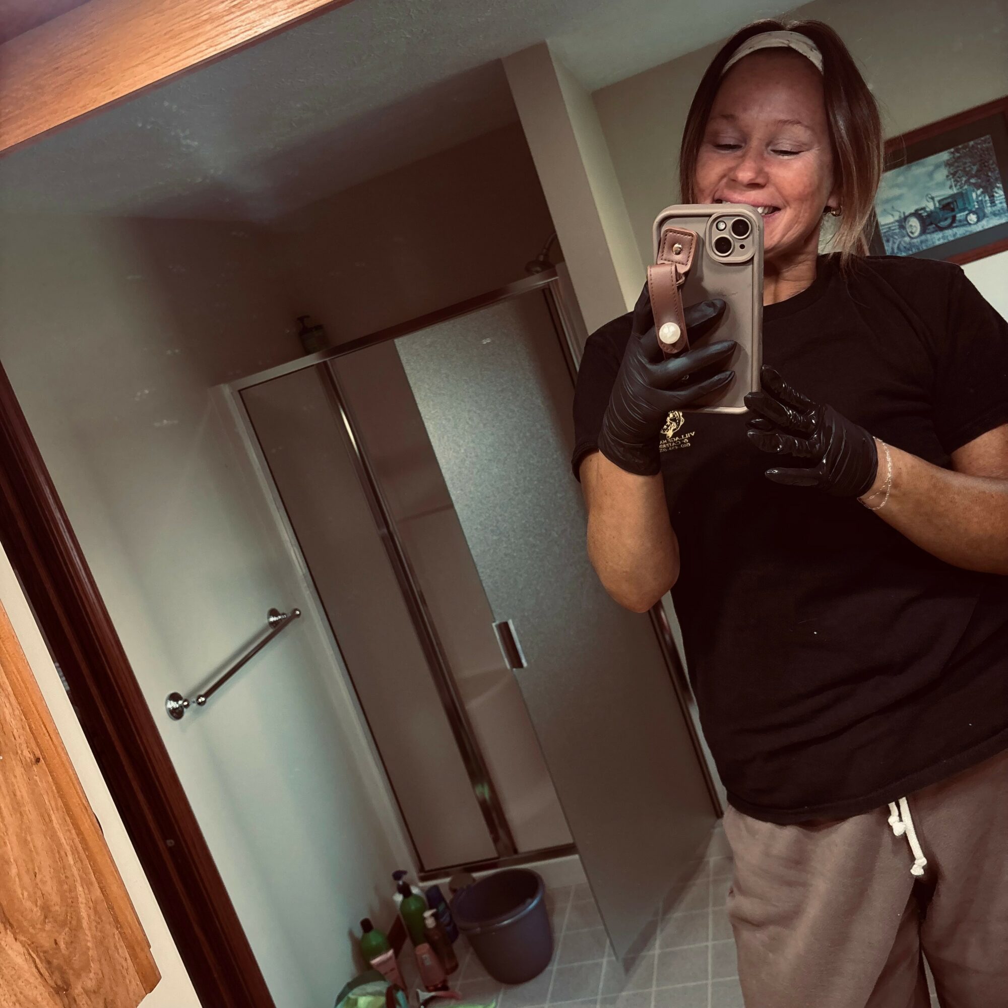Smiling woman taking a mirror selfie in a bathroom, wearing black gloves and a black shirt, with a shower and towel bar in background.