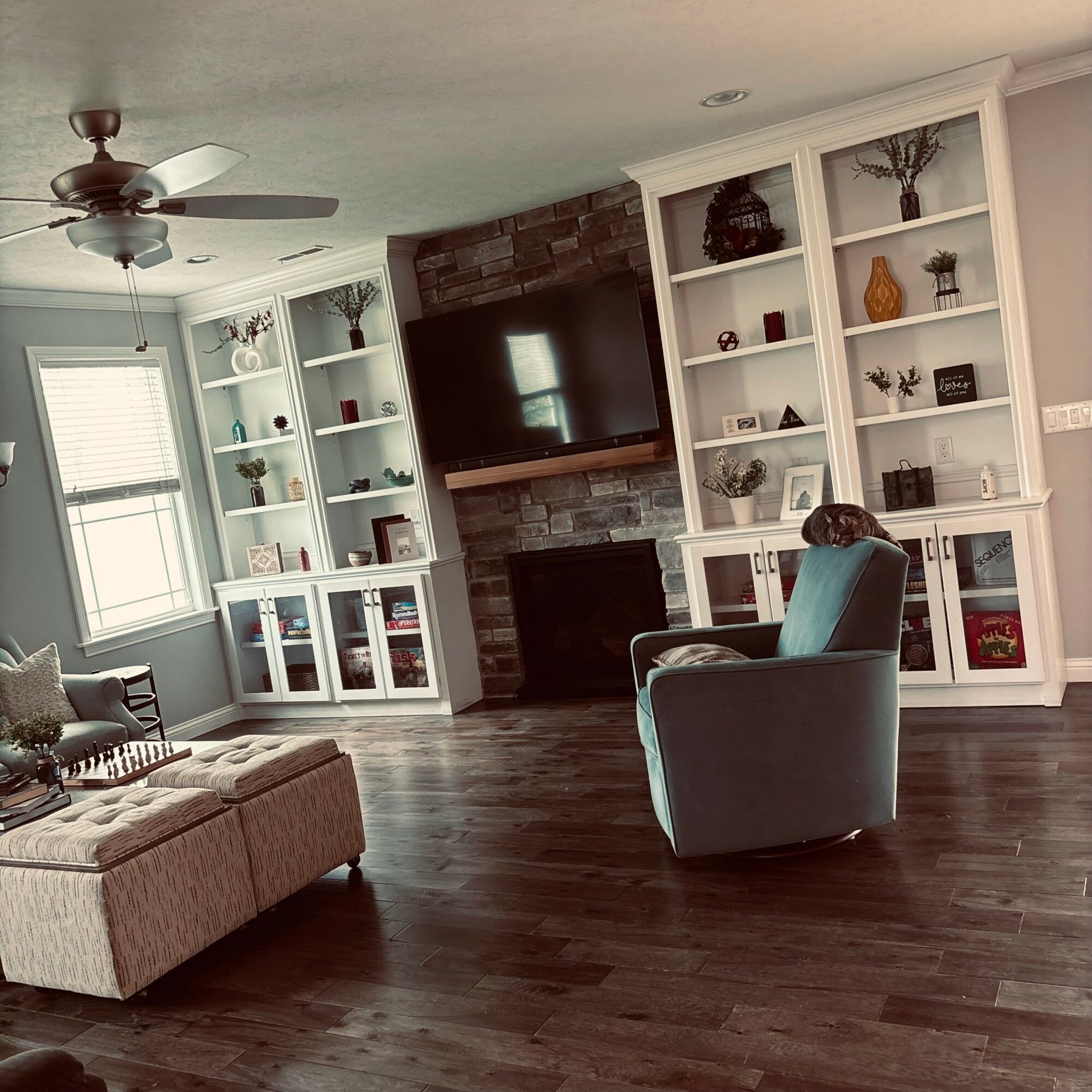 Living room with a ceiling fan, large window, built-in shelves, a TV above a fireplace, and furniture including an armchair and ottoman.
