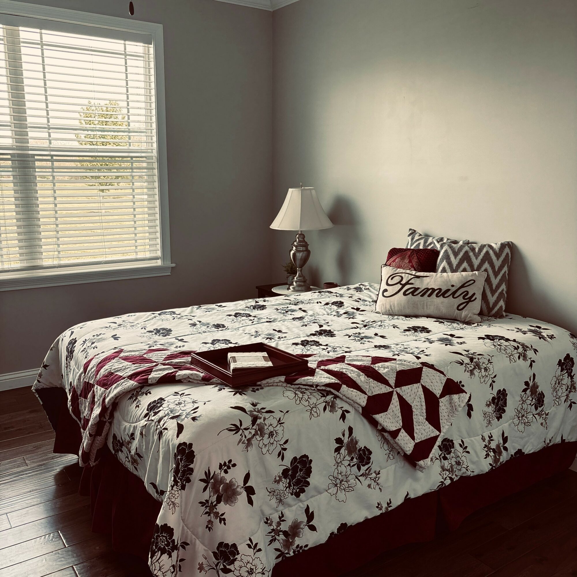 Bedroom with a bed, decorative pillows, a lamp, and a window with blinds, sunlight streaming in, wooden floor.