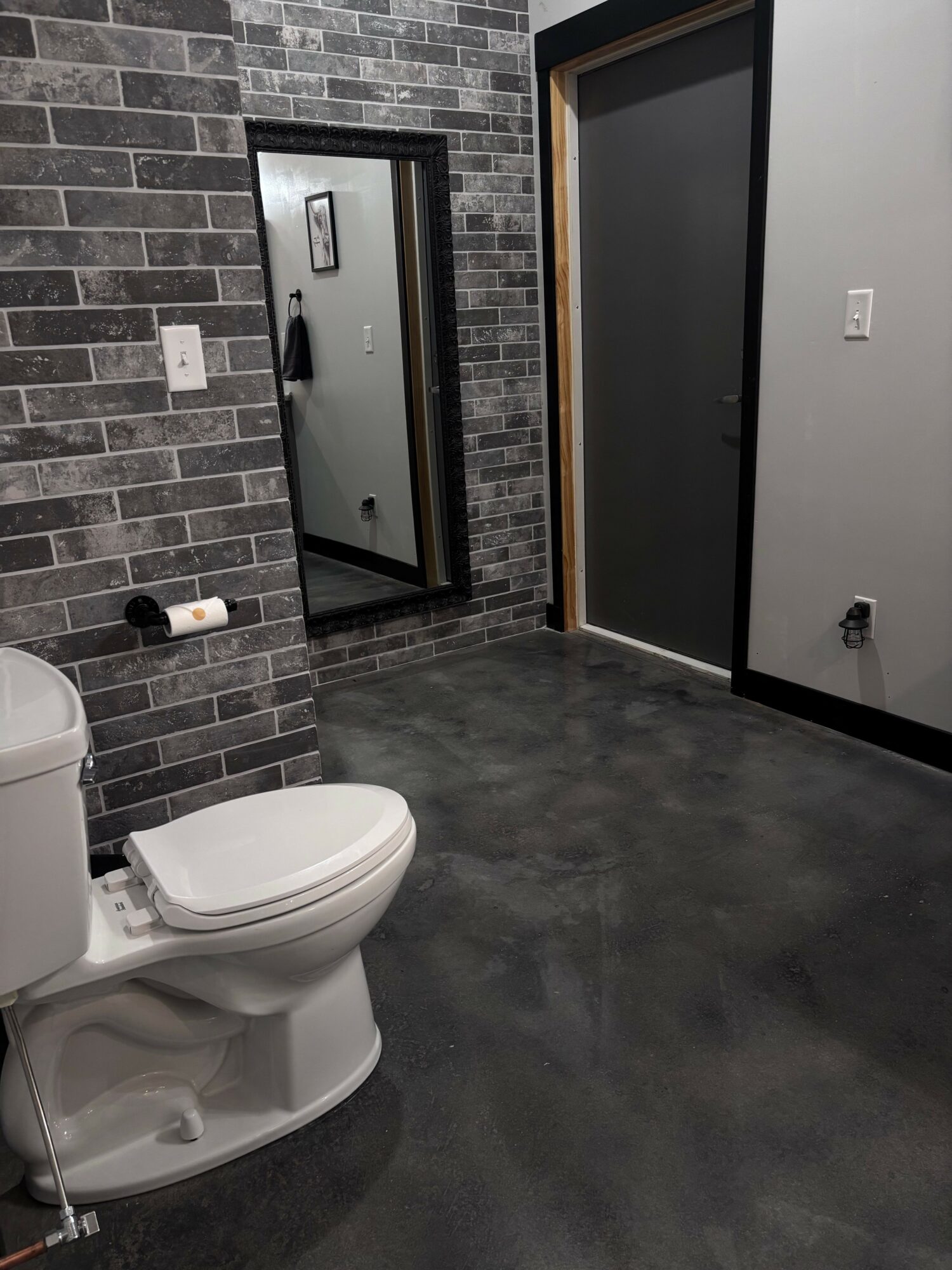 Toilet in a room with brick-patterned wall and dark floor, door and mirror visible in the background.