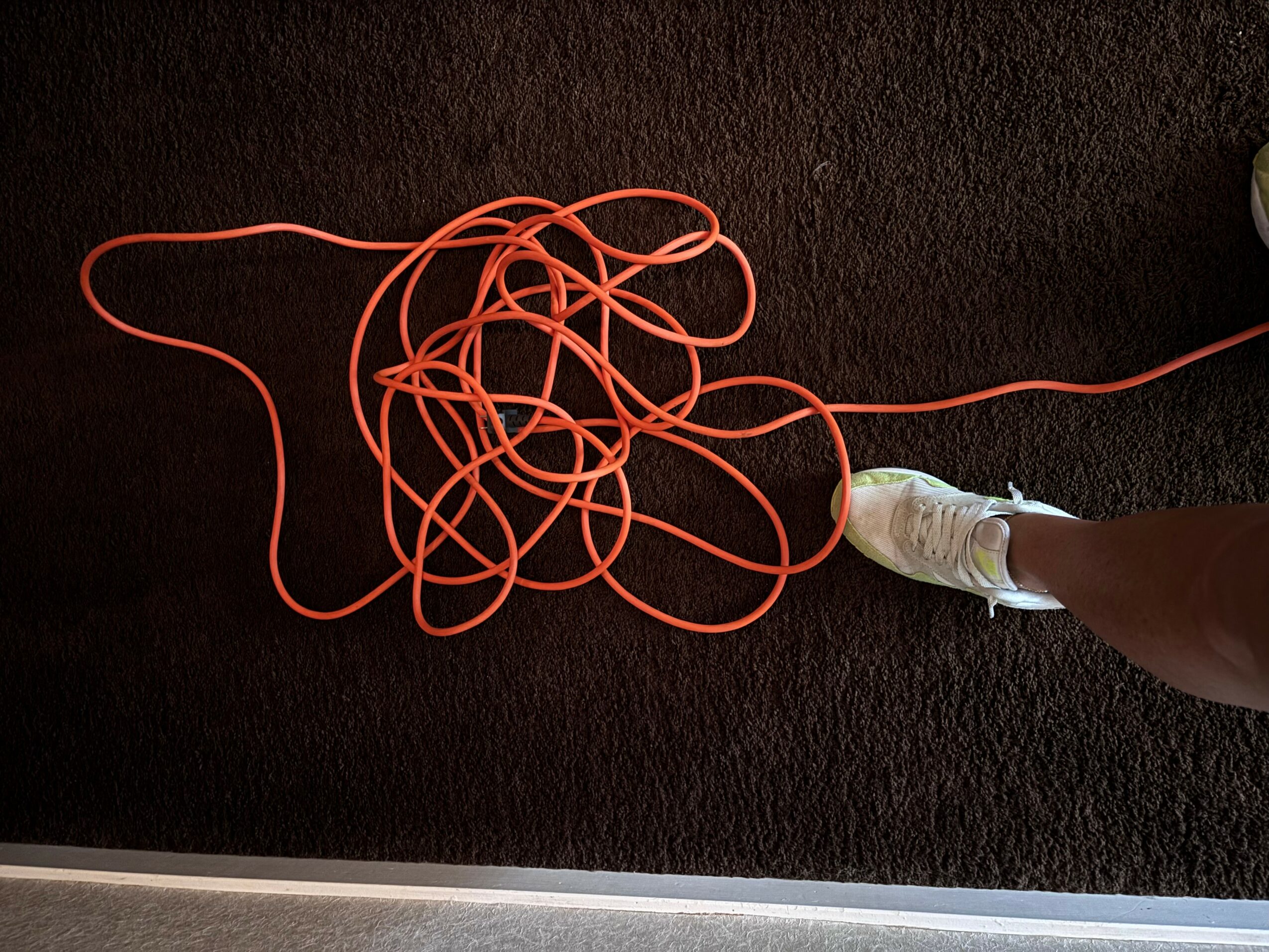 Person wearing a white shoe standing on dark carpet with tangled orange cord on the floor.