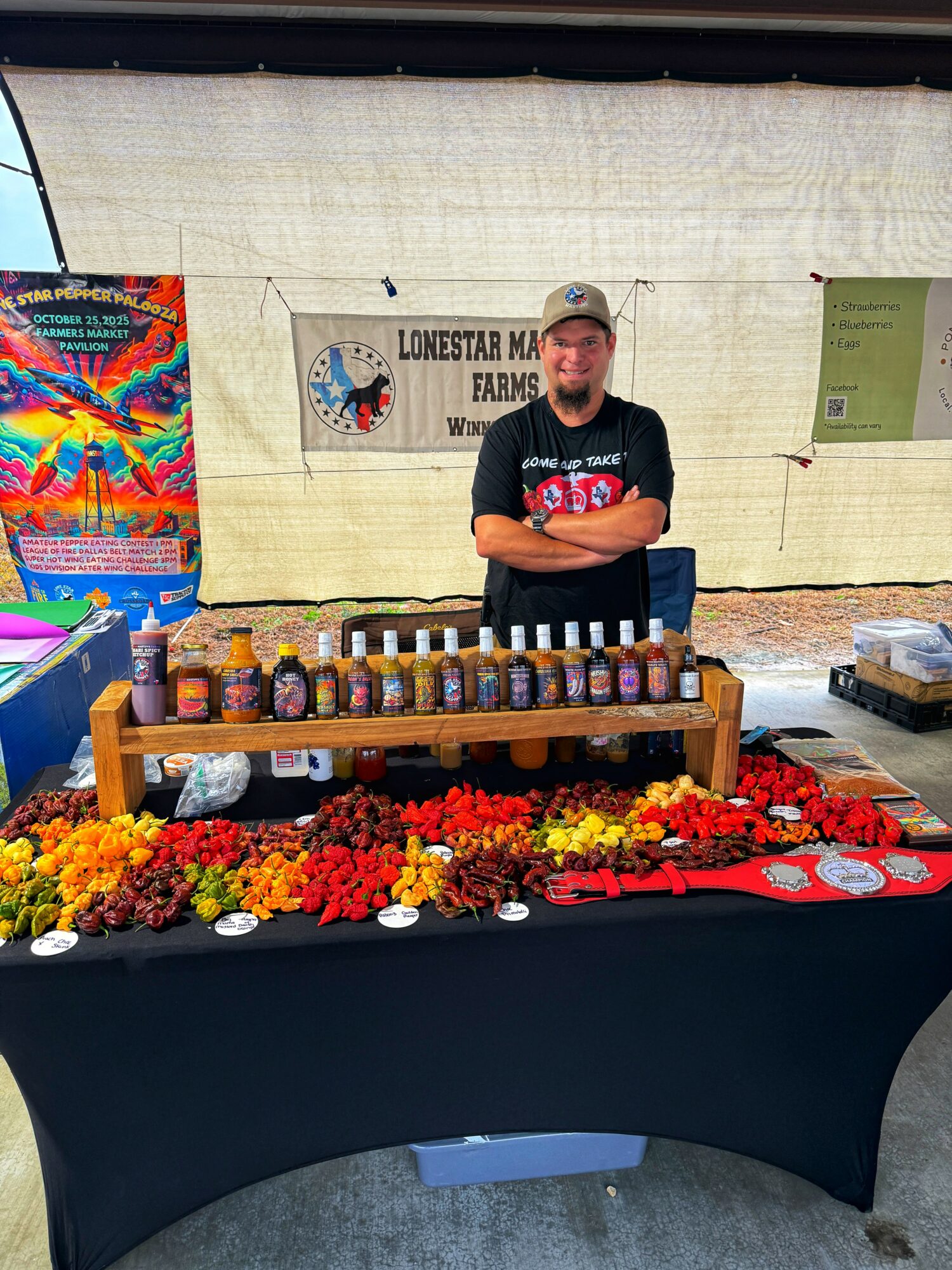 Man standing behind a table with jars and flowers at an outdoor market stall, smiling with arms crossed.