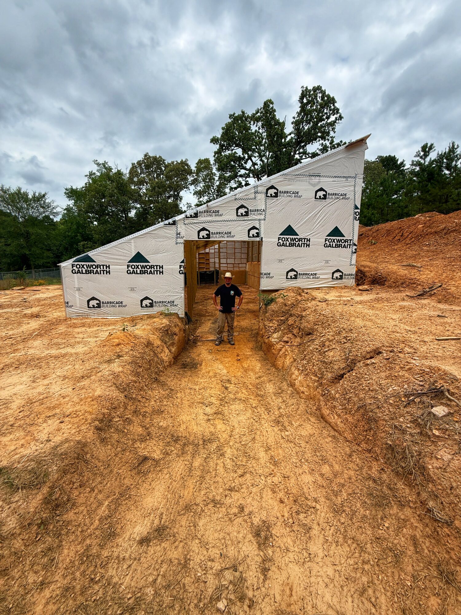 Person standing in front of a partially constructed building with a wooden door, on a dirt path, with trees and cloudy sky in background.