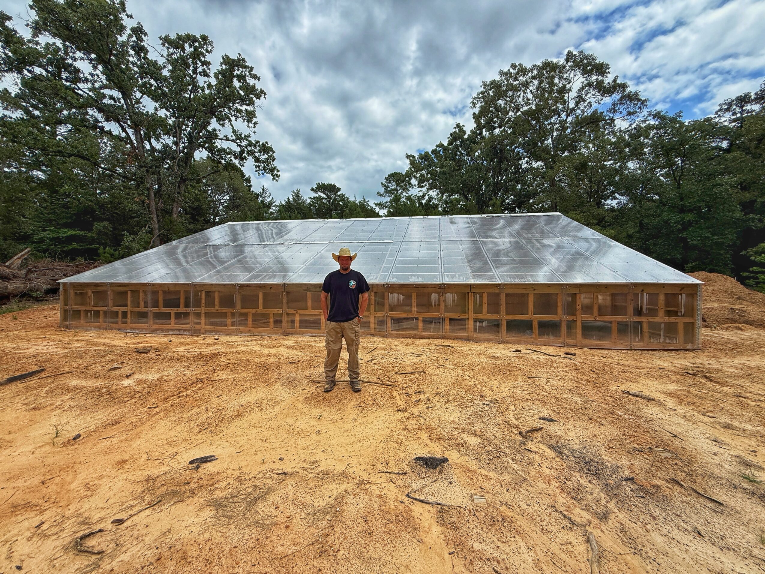 Person standing in front of a large building under construction with a metal roof, surrounded by trees and dirt ground.