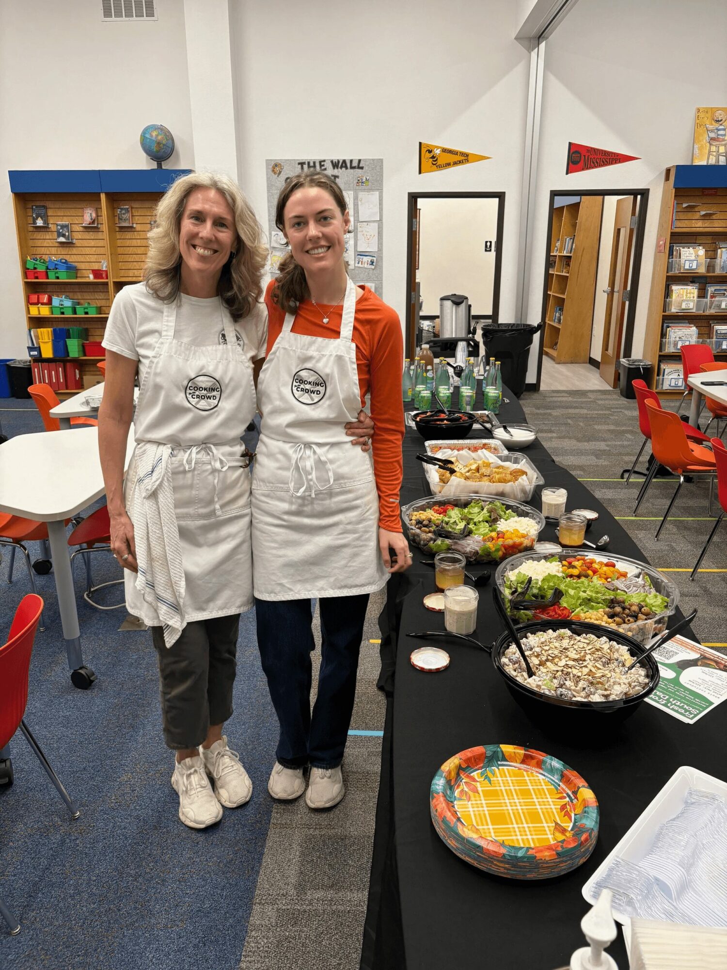 Two women wearing aprons stand behind a table with food in a room with colorful chairs and shelves.