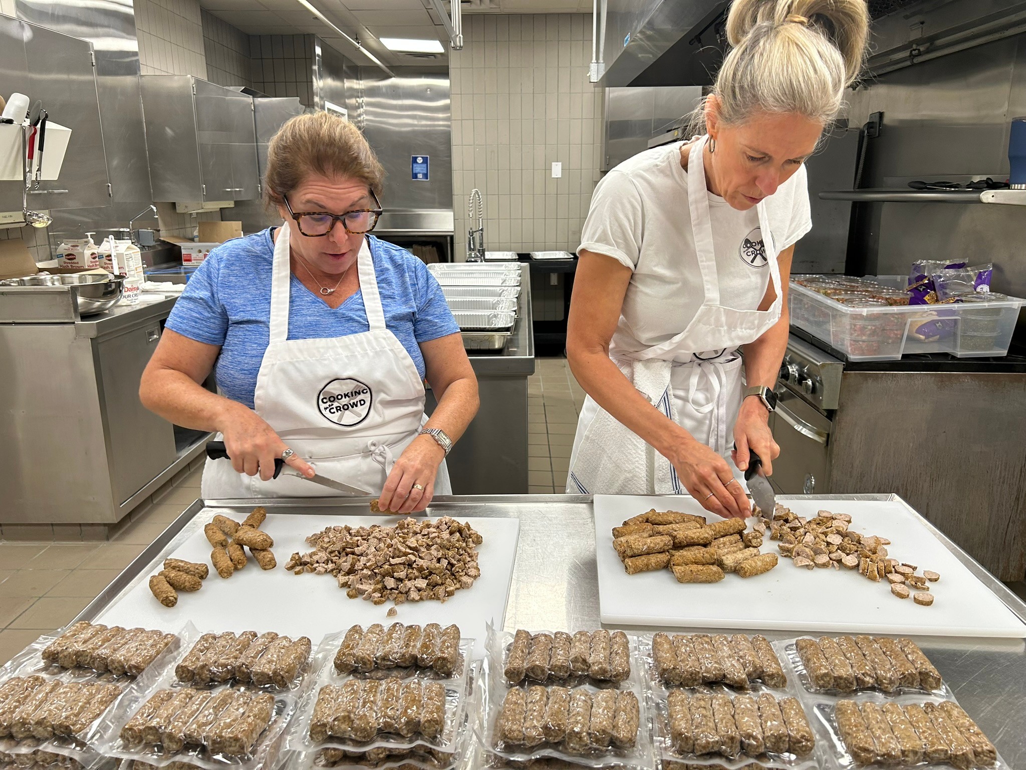 Two women in a commercial kitchen prepare food, with trays of cooked items in front of them.