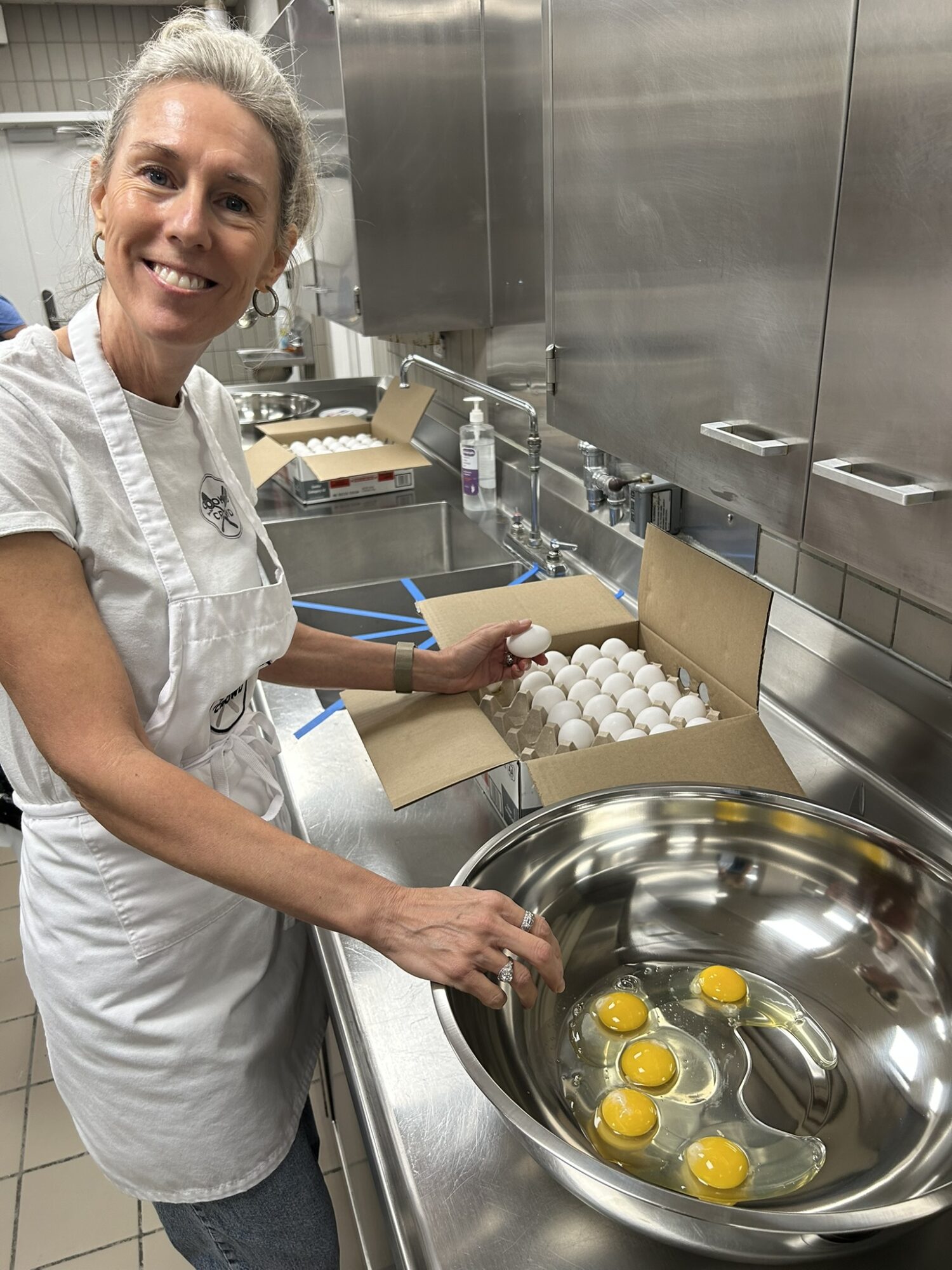 Smiling woman in white apron cracking eggs into a large metal bowl in a commercial kitchen.