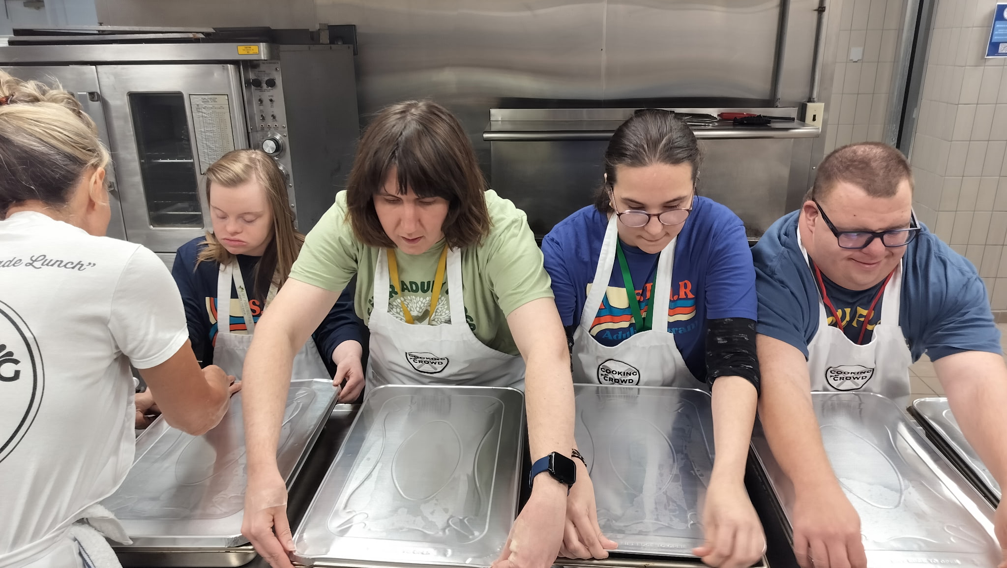 Four people wearing aprons work together in a kitchen, preparing food at a stainless steel counter.