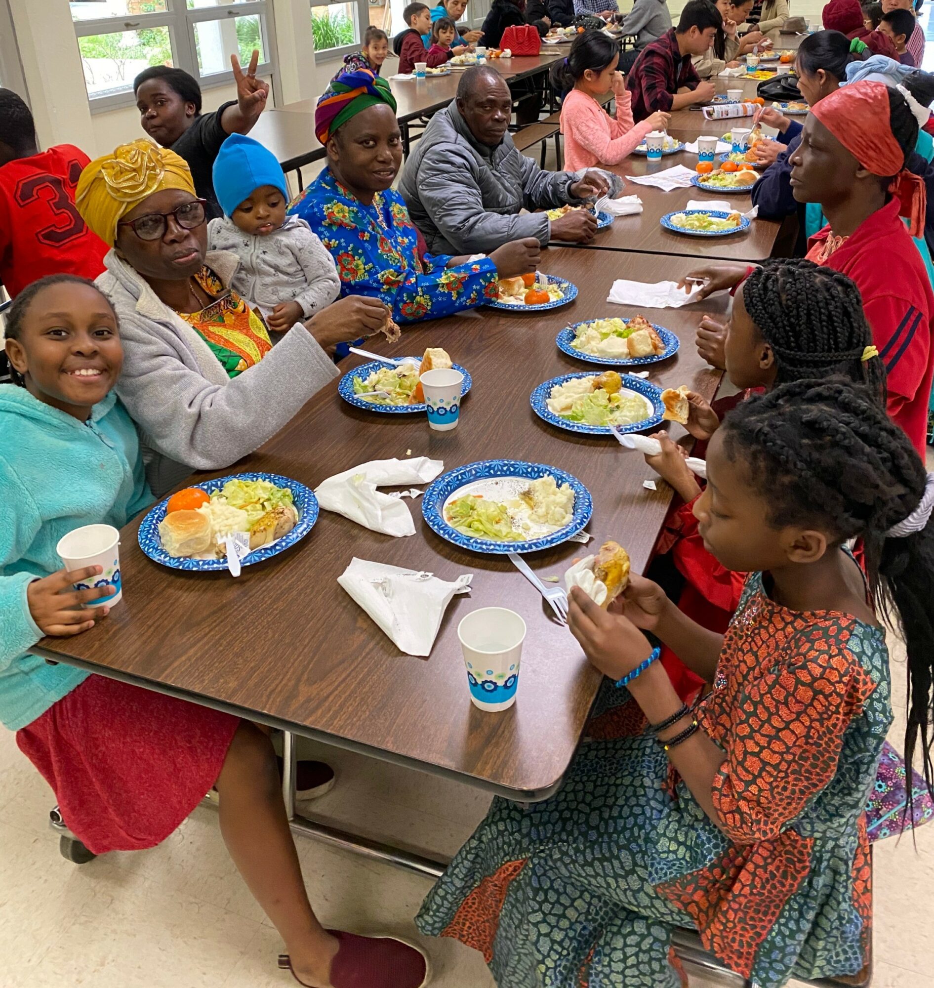 Group of diverse people, including children and adults, sitting at a table with food, smiling, in a bright room.