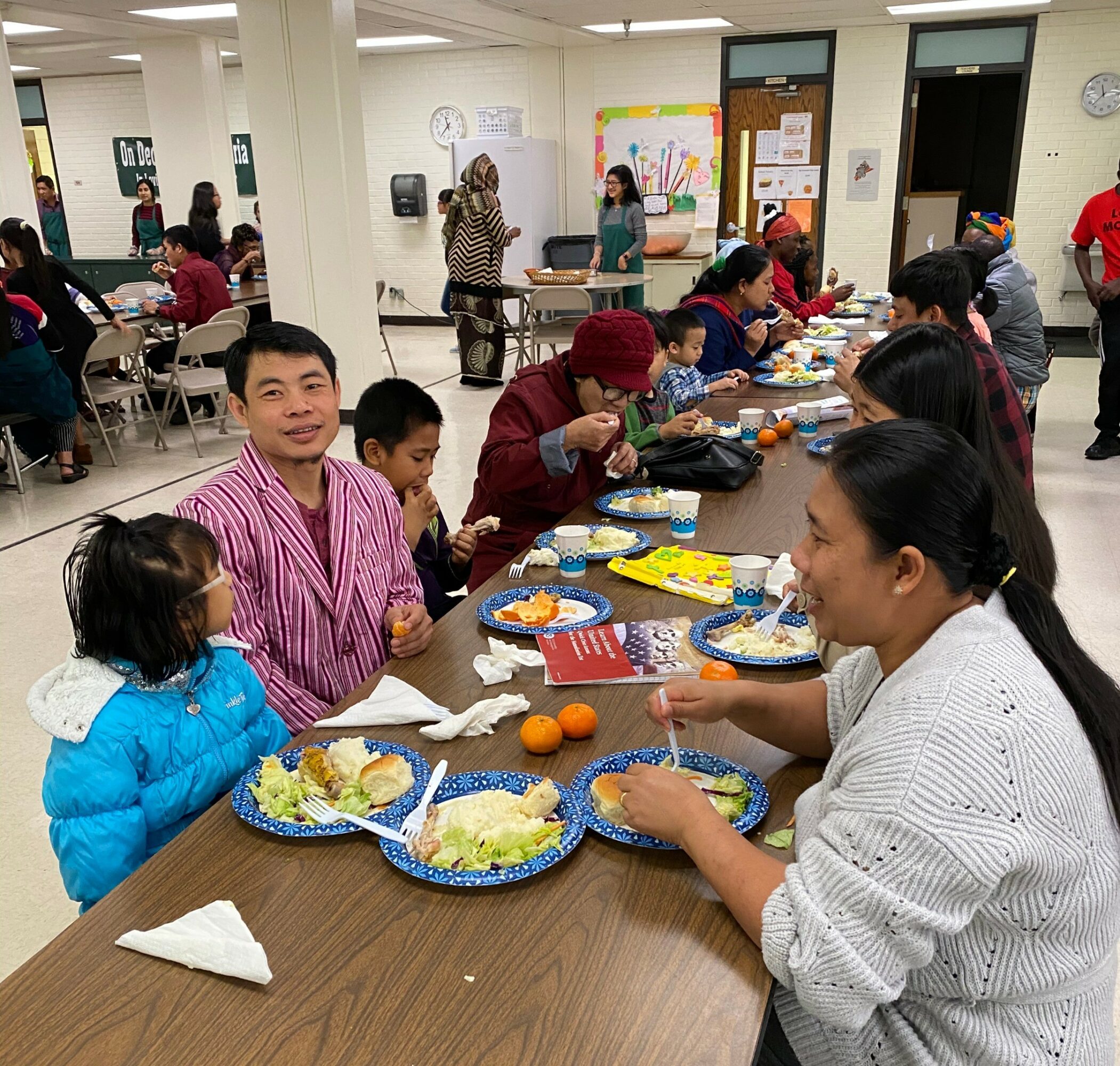 People sitting at a long table eating food in a community room, with others in the background. Bright lighting and white walls.