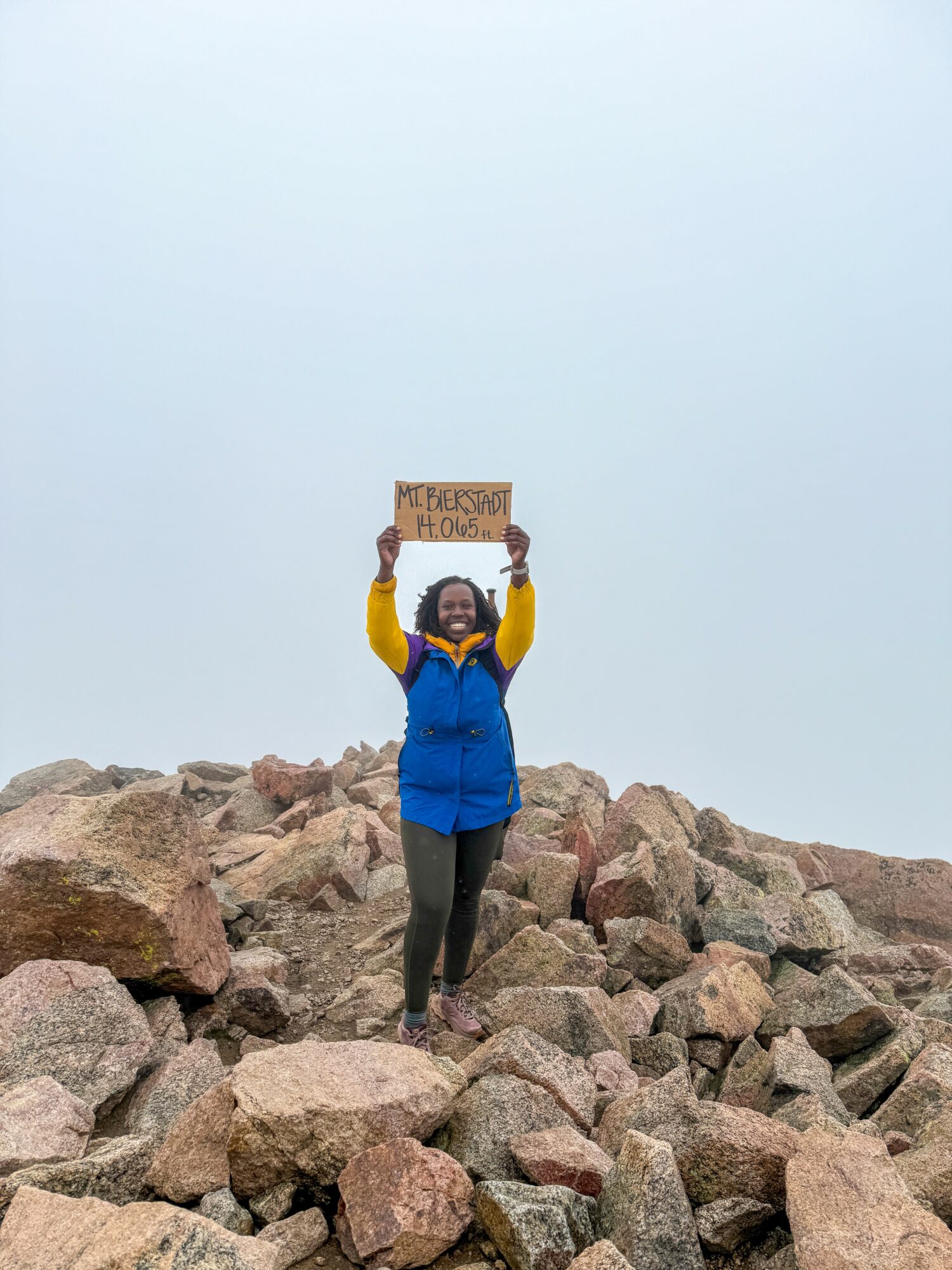 Person standing on rocks holding a sign above their head, smiling, with a cloudy sky background.