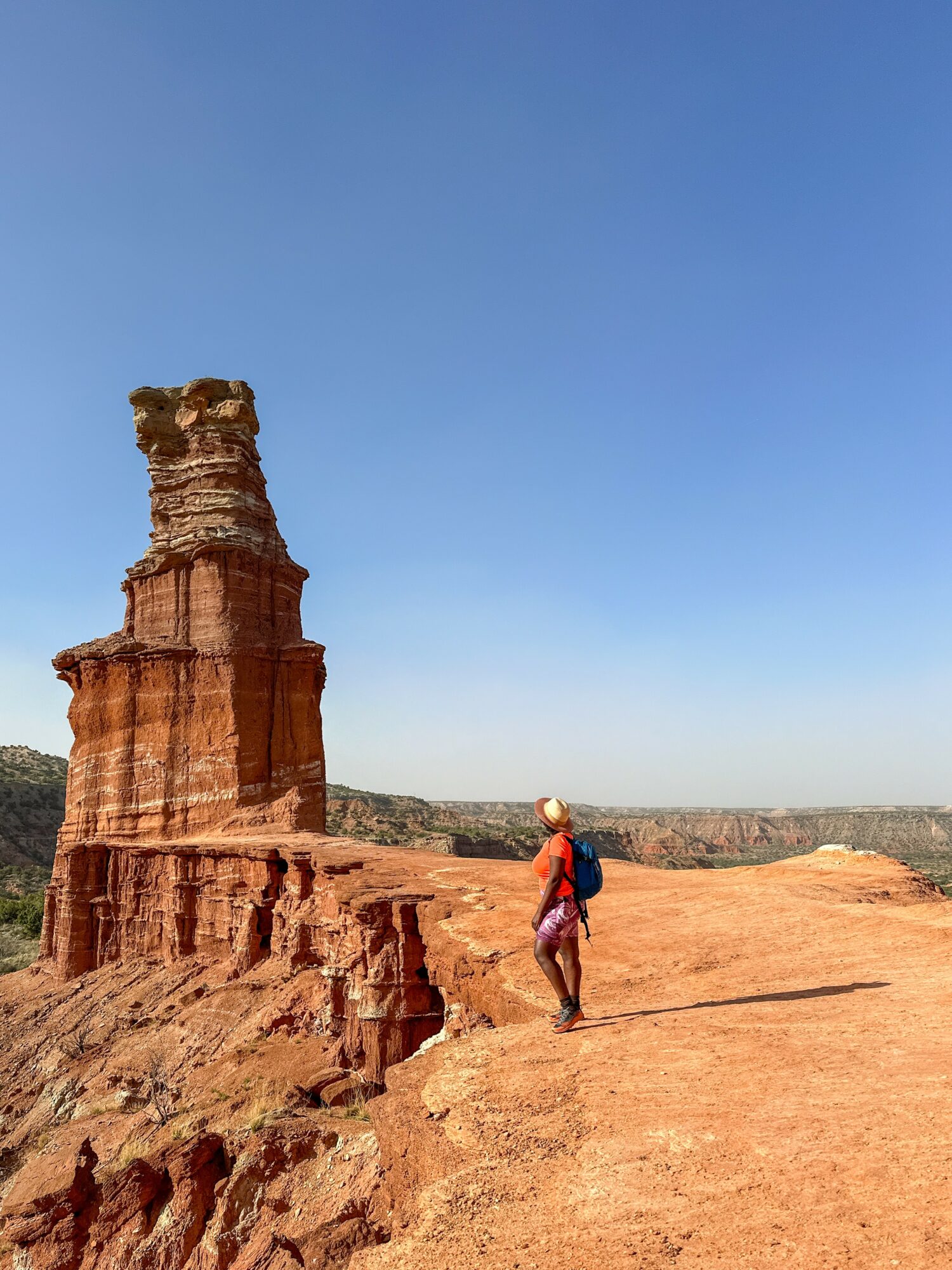Person walking on desert terrain near tall rock formation under clear blue sky.
