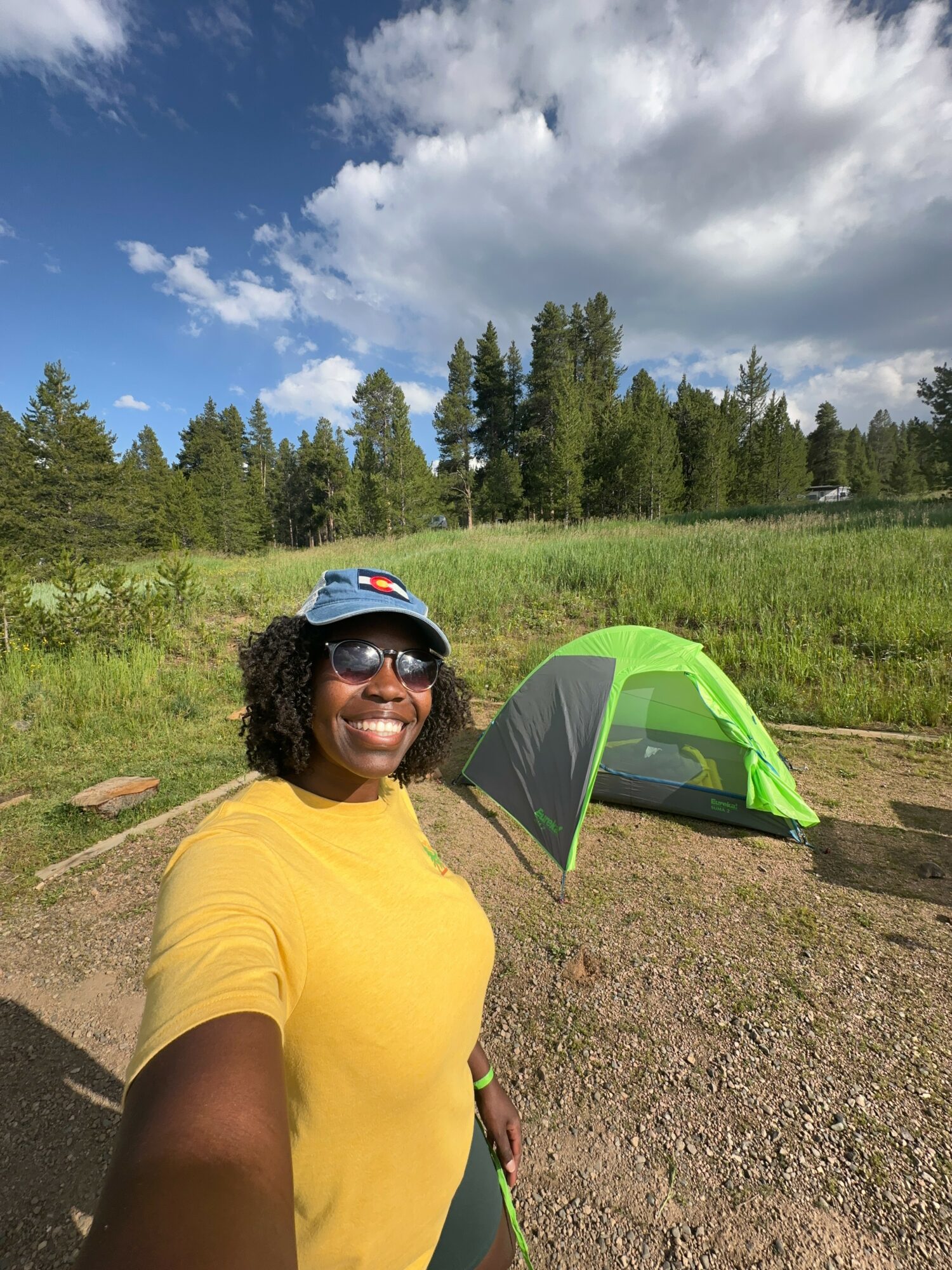 Person wearing sunglasses and a blue cap takes a selfie outdoors with a green tent and trees in the background.