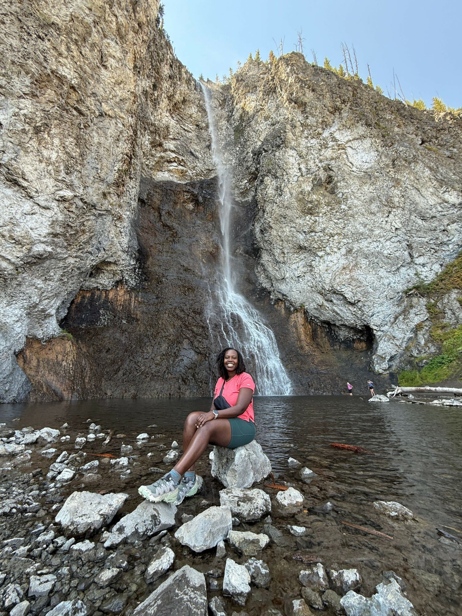 Person crouching near a waterfall in a rocky stream, wearing a pink top and shorts, with water flowing behind.