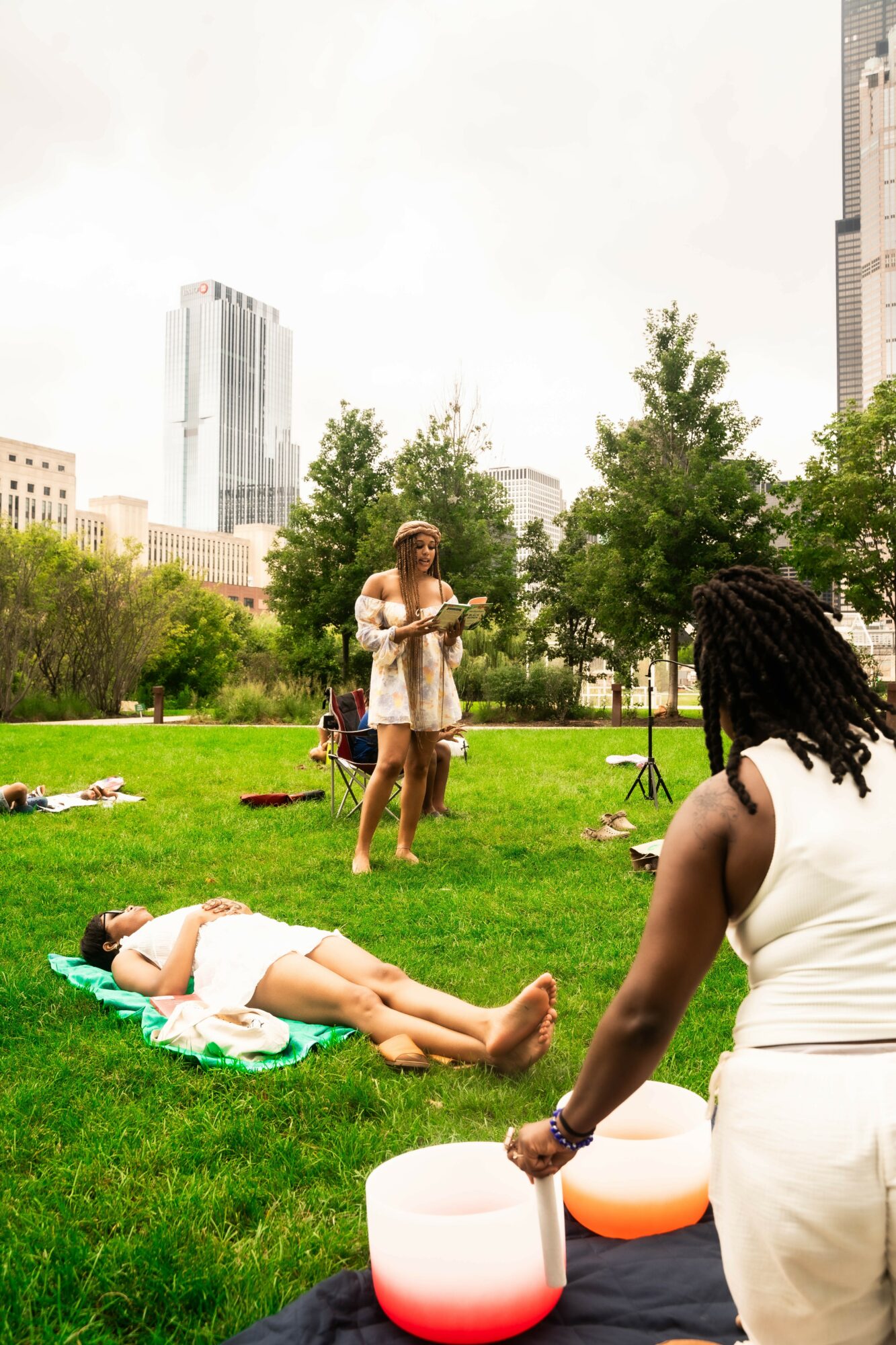 People relaxing on grass in a park with city buildings in background, woman lying on towel, others standing and sitting, trees present.