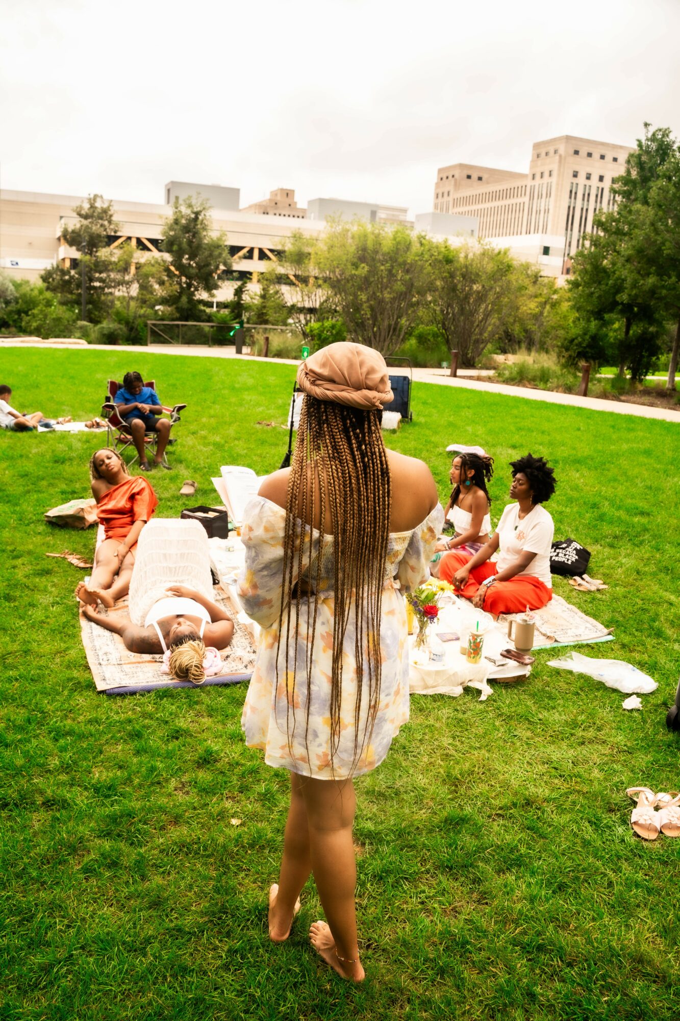 Group of children and a woman on a picnic blanket in a park with trees and buildings in the background.