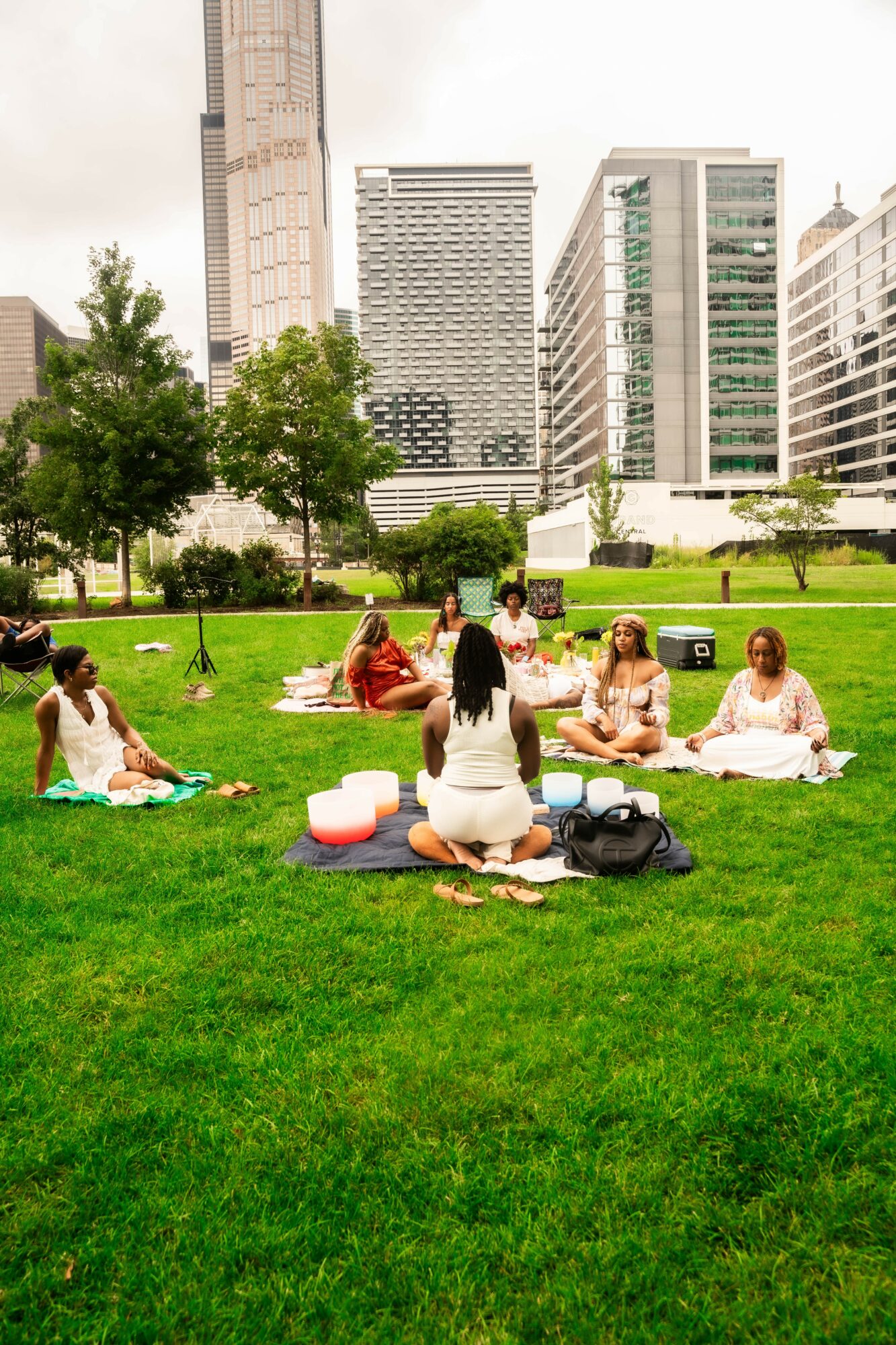 Group of people practicing yoga outdoors on a grassy area in a city park with tall buildings in the background.
