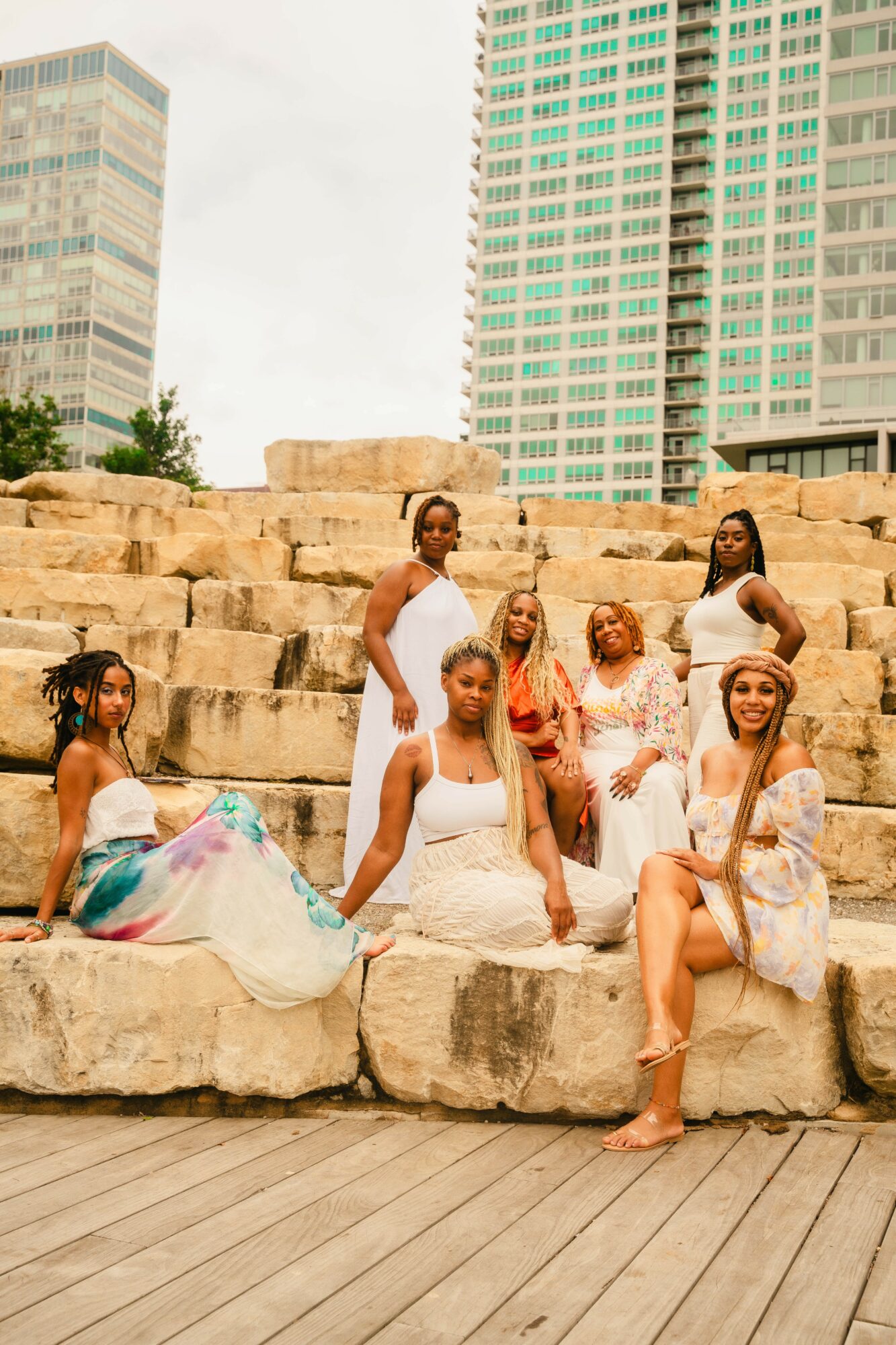 Seven women in white dresses sitting and standing on stone steps outdoors with tall buildings in background.