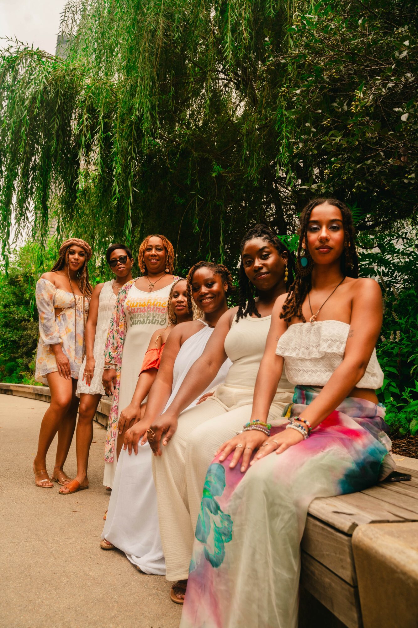 Six women sitting and standing outdoors with green trees and hanging branches in the background.