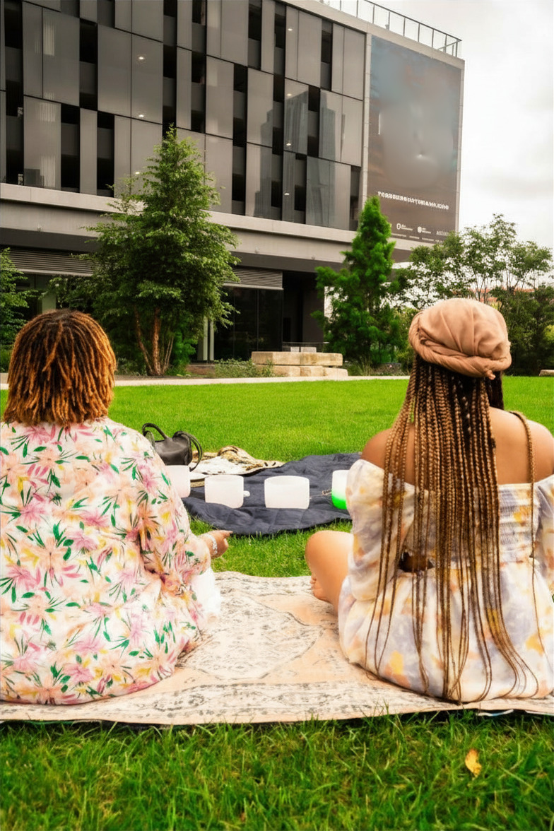 Two women sit on a blanket outdoors with a modern building and trees in the background.