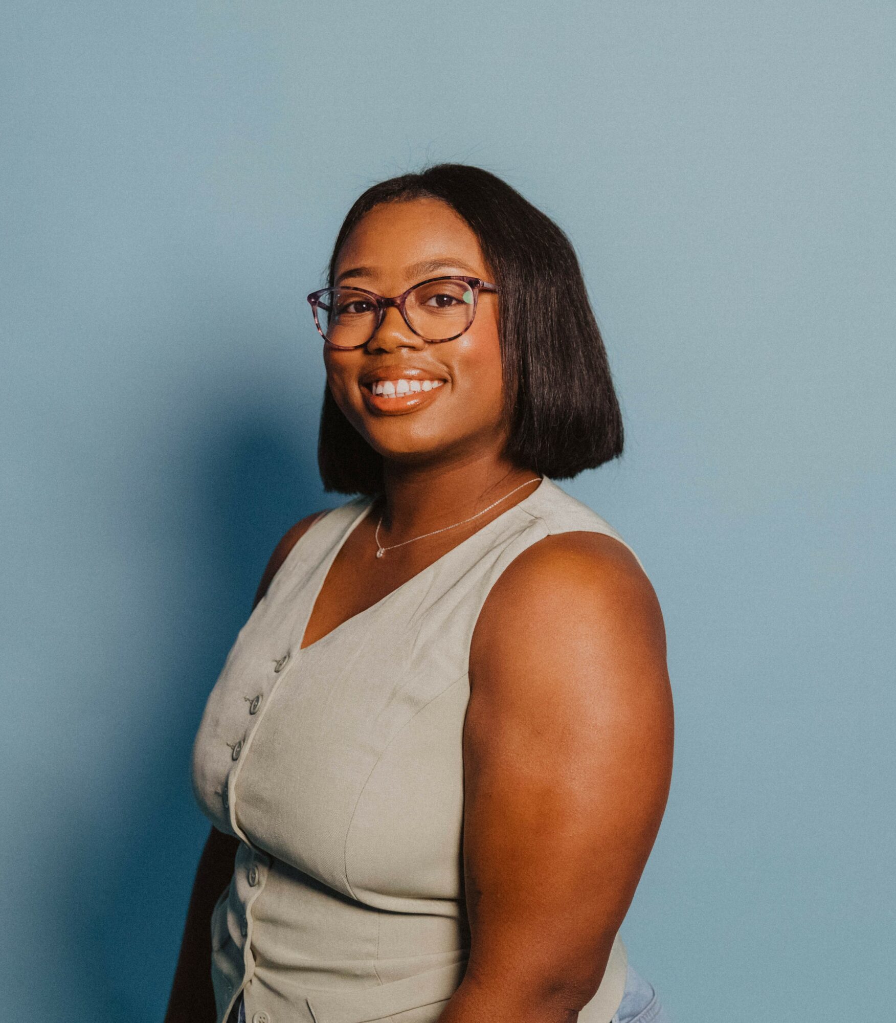 Smiling woman with glasses and shoulder-length hair, wearing a sleeveless light-colored top, standing against a blue background.