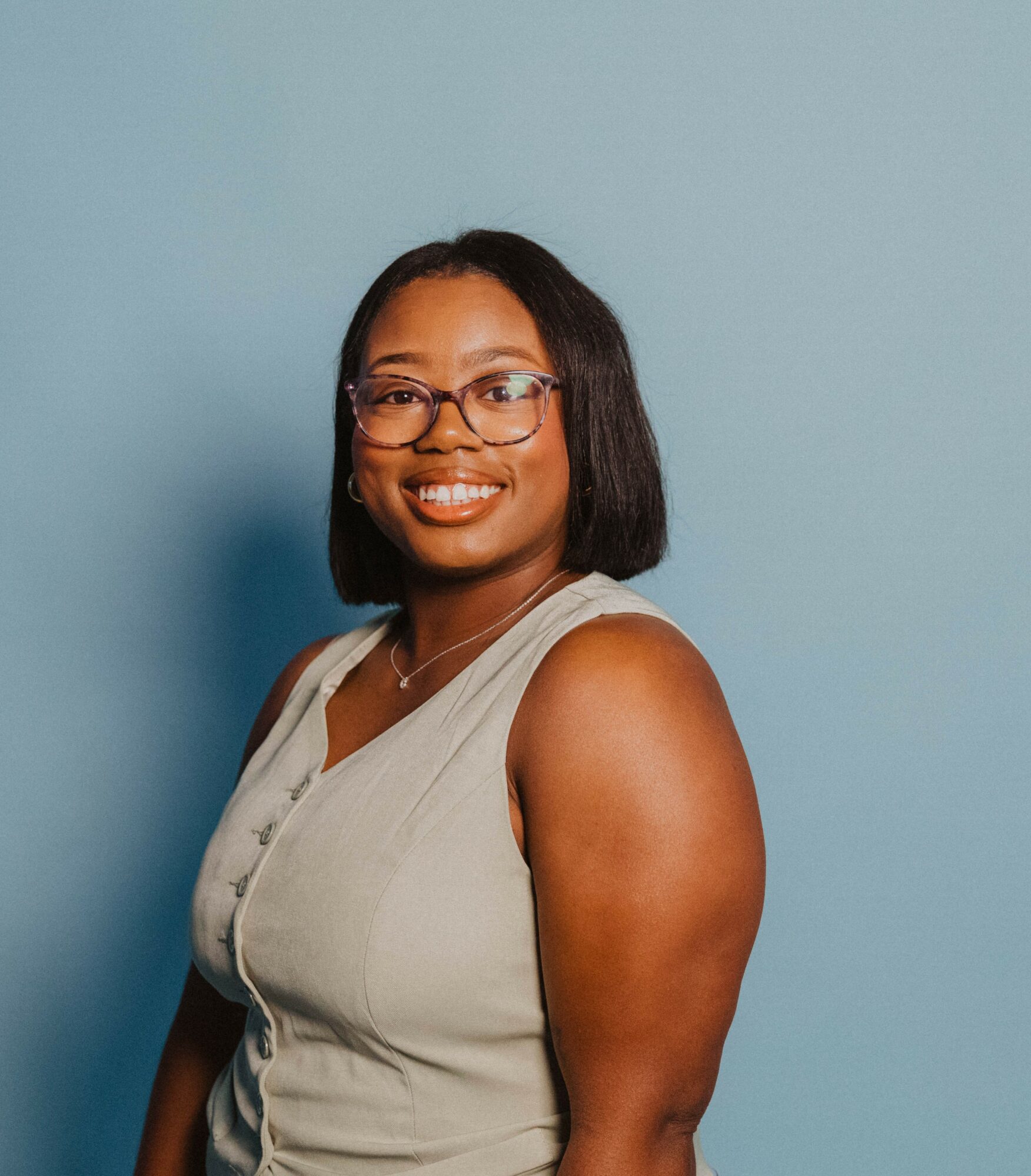 Smiling woman with glasses and shoulder-length hair, wearing a sleeveless top, standing against a light blue background.